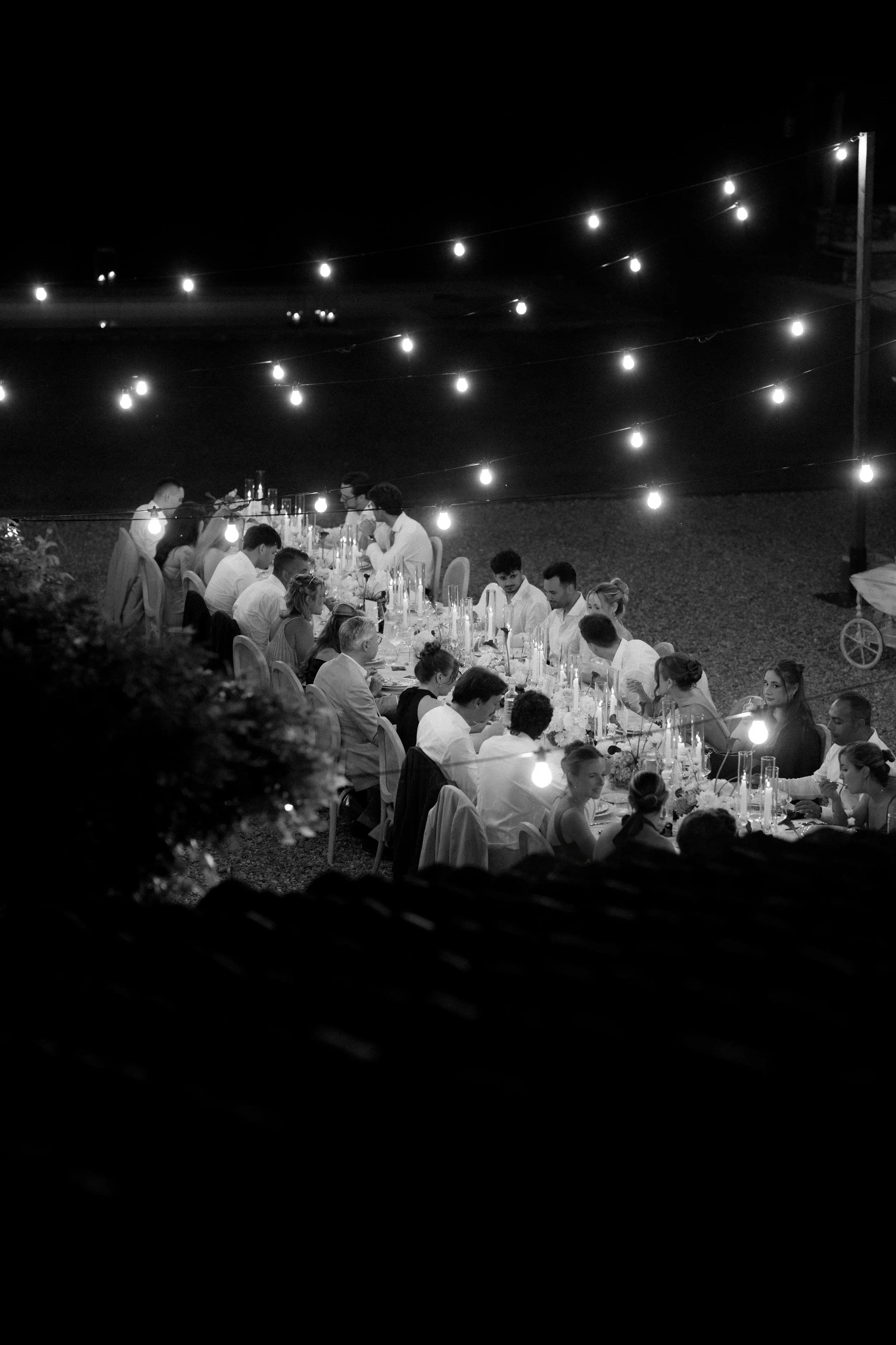 Eine Gruppe von Menschen sitzt an einem langen Tisch, der für eine Abendveranstaltung gedeckt ist, unter Lichterketten bei Nacht.