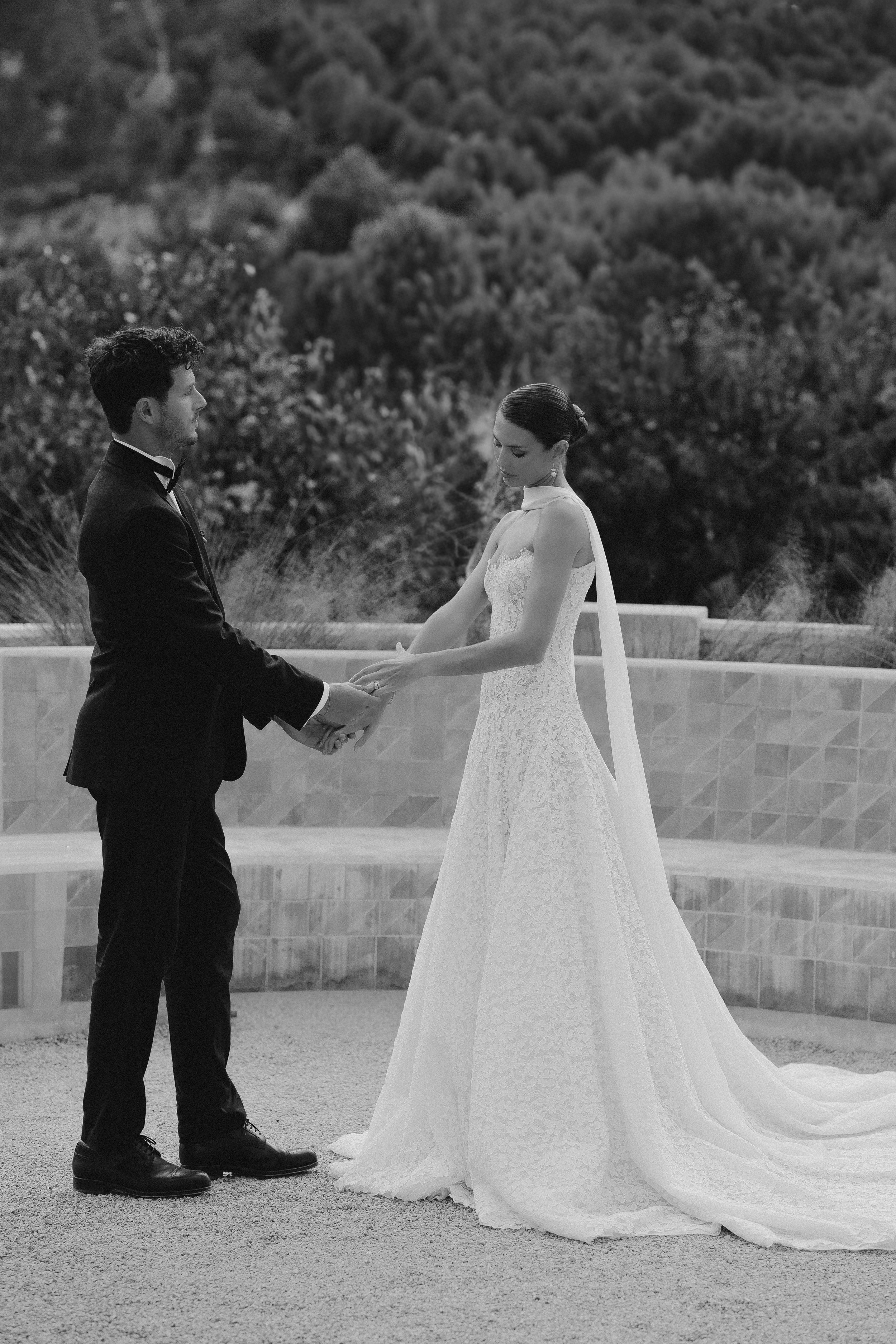 Ein Brautpaar hält sich bei ihrer Hochzeit im Freien die Hände und schaut sich an, im Hintergrund grüne Bäume.