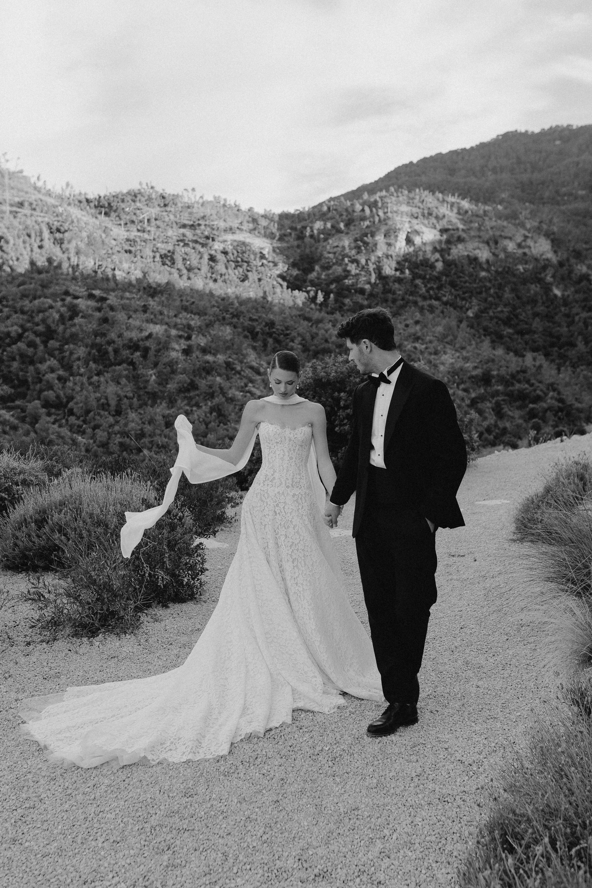 Ein Brautpaar in formeller Kleidung hält Händchen auf einem Sandpfad in einer Landschaft mit Hügeln und Büschen, schwarz-weiße Aufnahme.