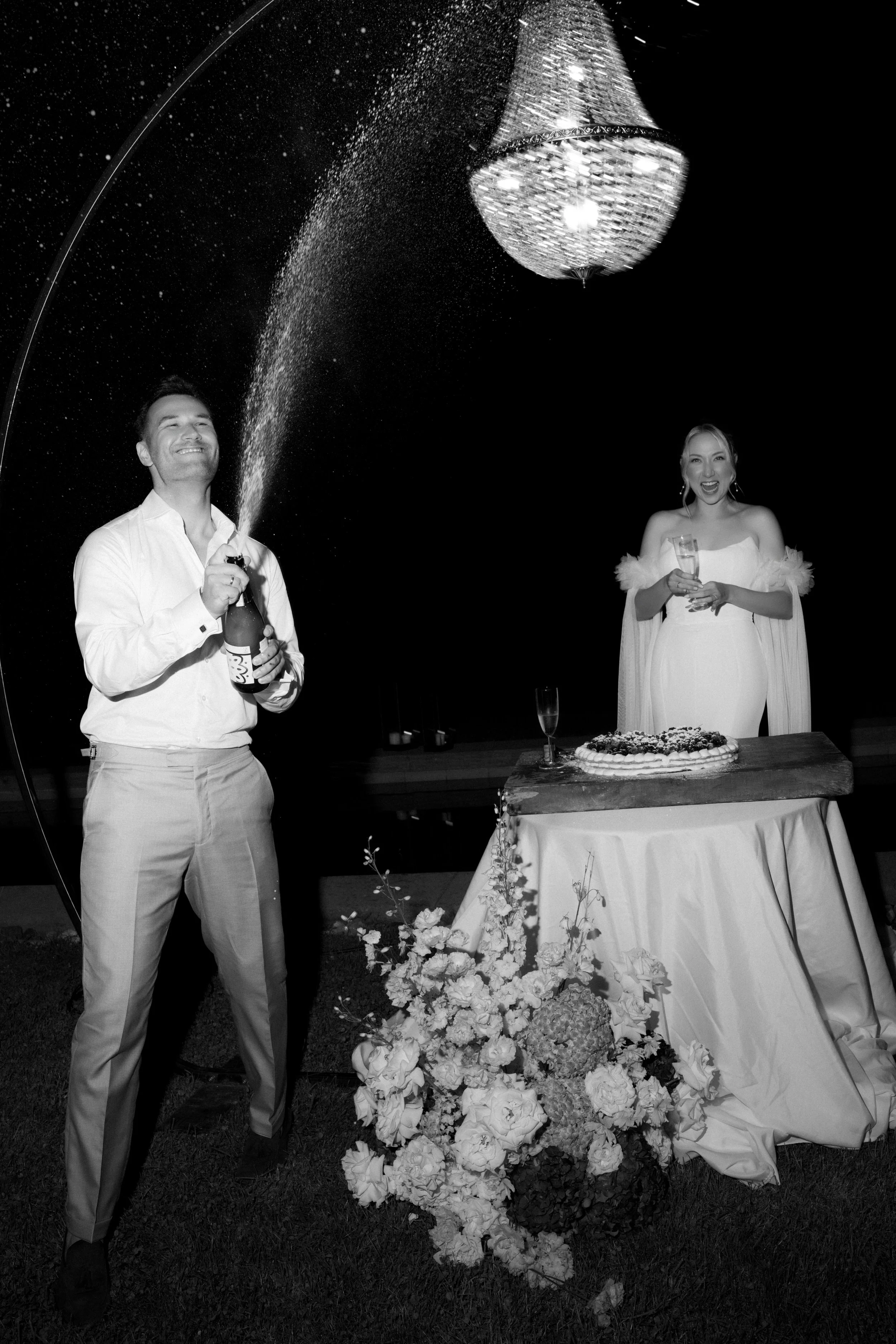 Ein lachendes Paar bei einer Hochzeit, das Feuerwerkskorken und Champagner trinkt, vor einem dekorierten Tisch mit einem Hochzeitskuchen und Blumen. Es ist Nacht, der Himmel ist dunkel, und eine große Lampe hängt über ihnen.