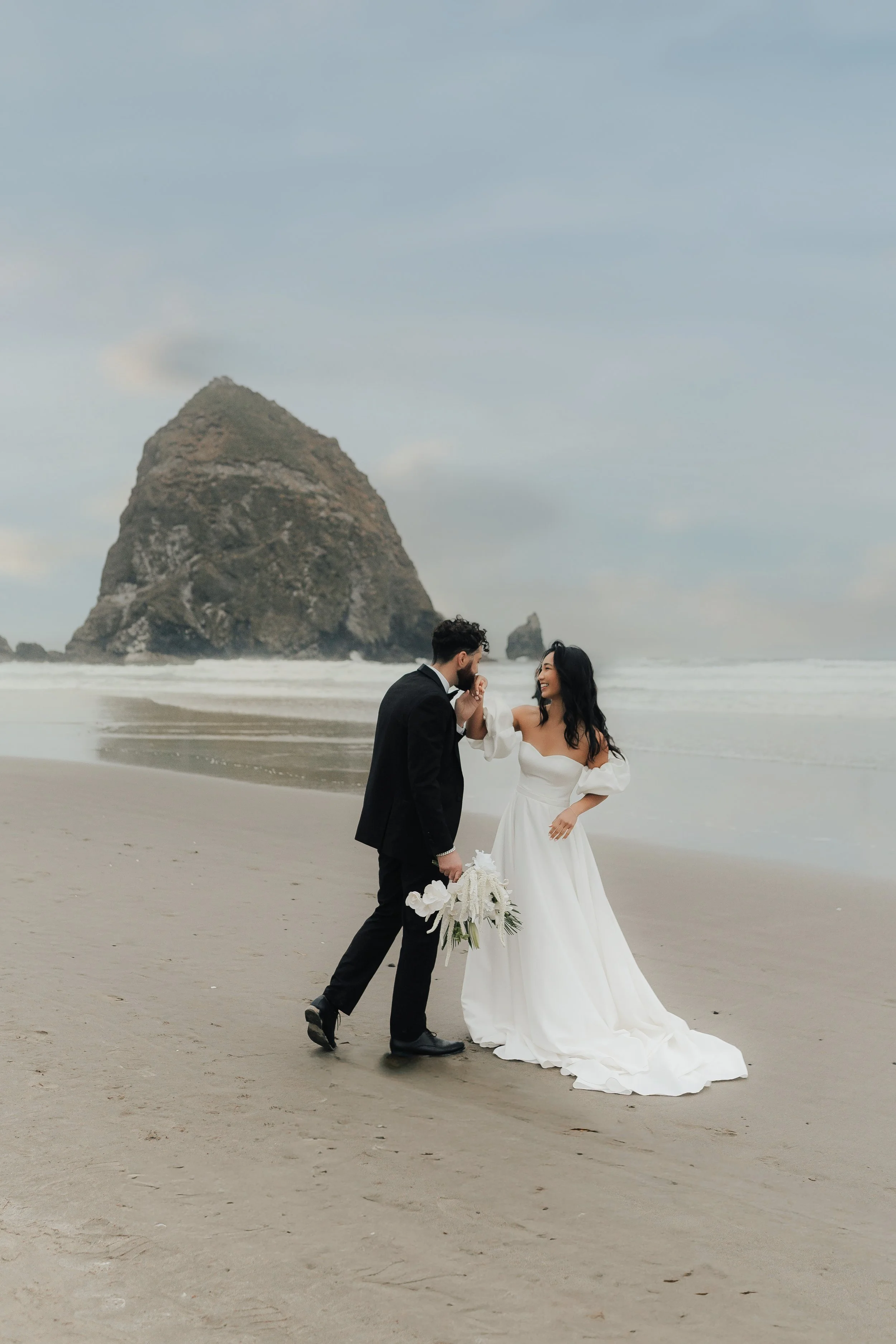 Ein Brautpaar in Hochzeitskleidung am Strand, im Hintergrund eine große Felsformation im Meer.