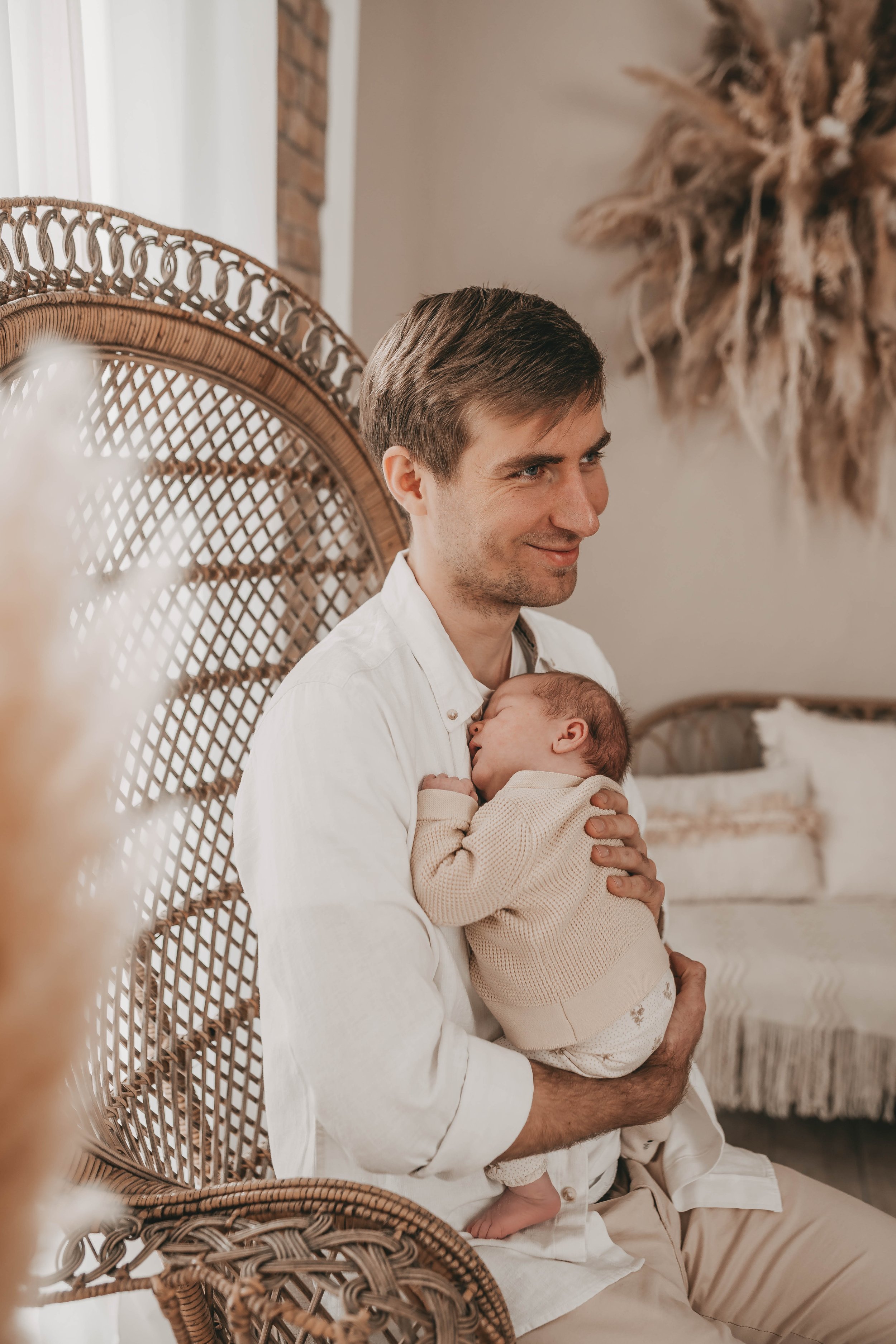 Mann hält schlafendes Baby im Arm, sitzt auf einem Rattanstuhl, gemütliche Inneneinrichtung im Hintergrund.