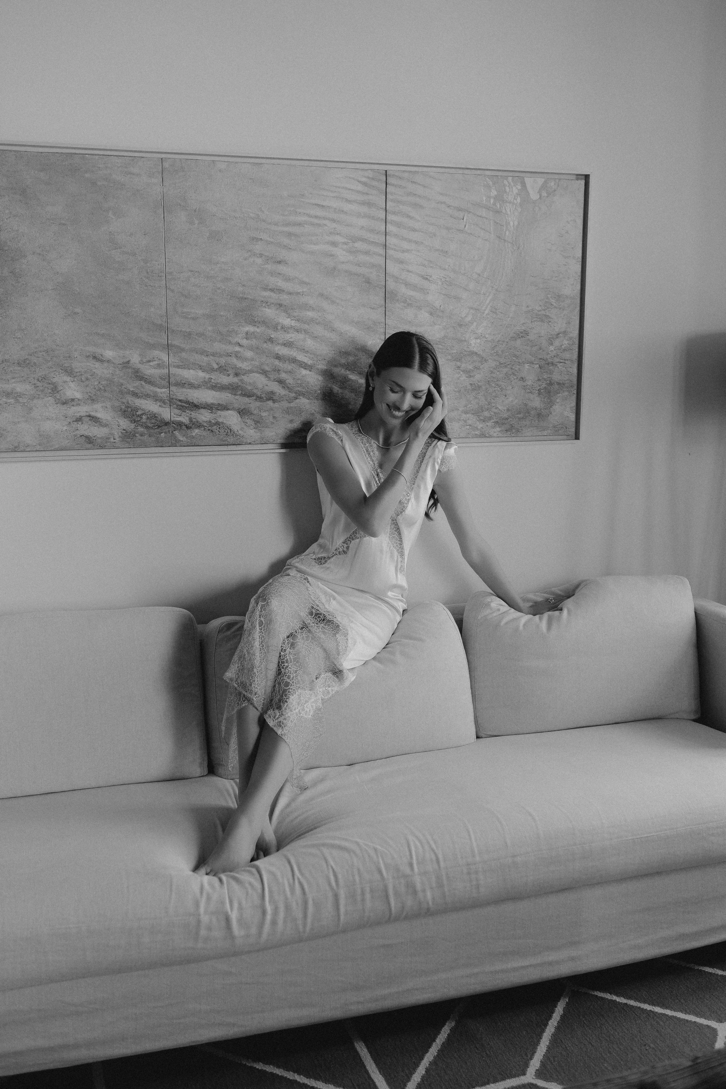 Frau sitzt lachend auf einem Sofa, berührt ihr Ohr, trägt ein langes, elegantes Kleid, im Hintergrund eine Wand mit Kunstwerk.