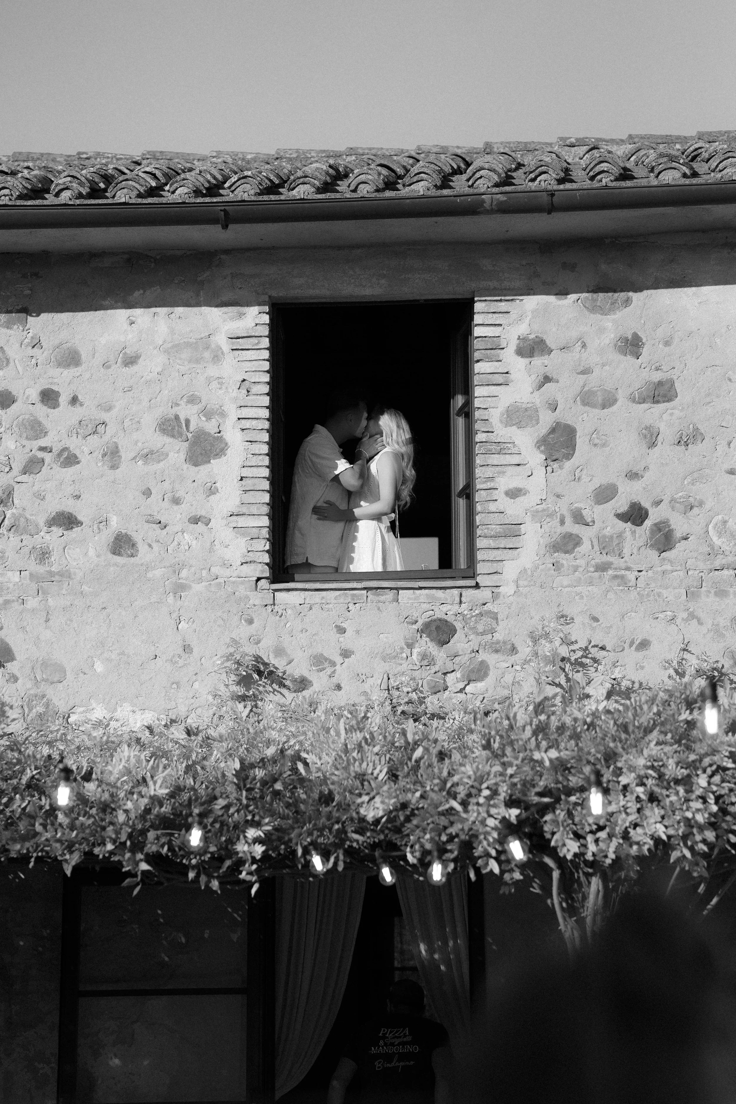 Ein schwarzer und weißer Blick durch ein Fenster in einer Steinwand auf ein Paar, das sich küsst, während es ans Fenster tritt, im Hintergrund eine mit Lichterketten geschmückte Kulisse.