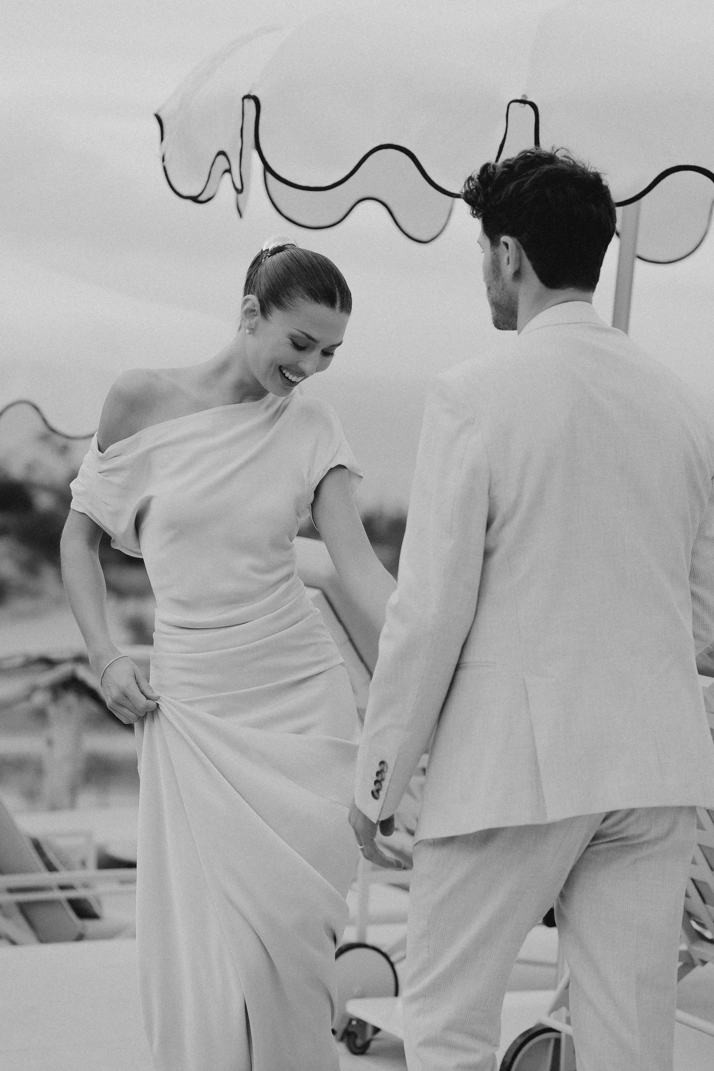 Ein Mann und eine Frau am Strand, sie lachen und halten Händchen, im Hintergrund Sonnenschirme und Liegestühle.