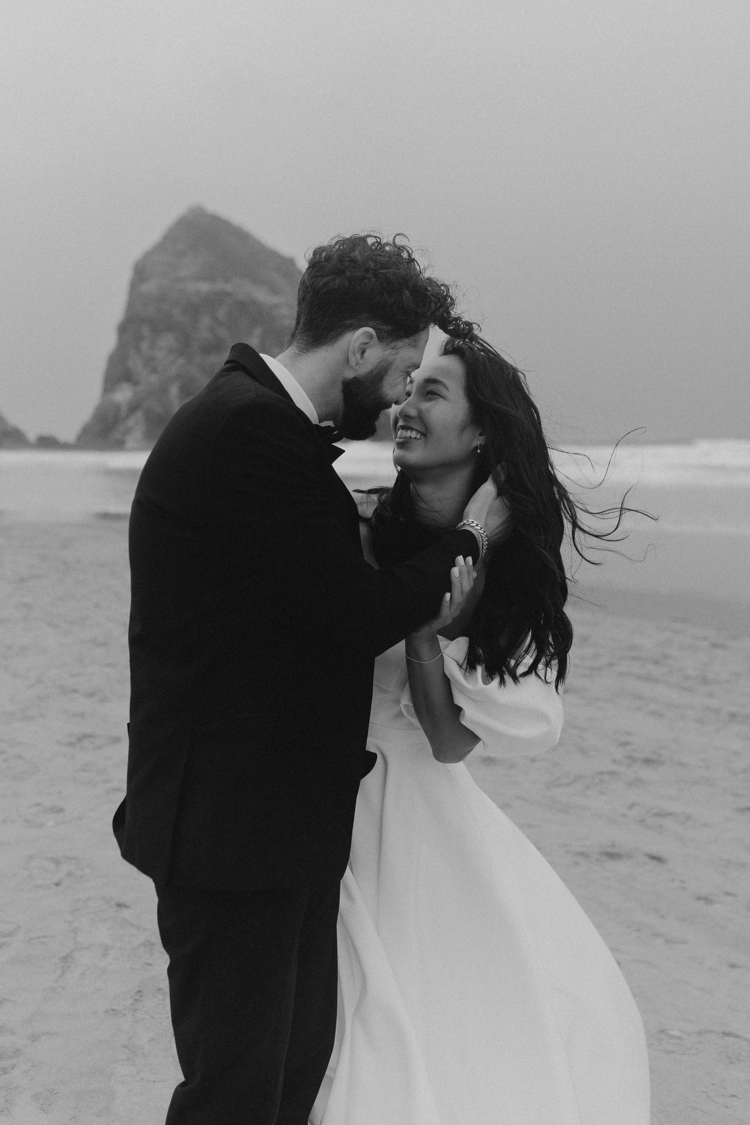 Ein glückliches Paar in formeller Kleidung, die sich an einem Strand umarmen und küssen, mit einer Felsenformation im Hintergrund, in Schwarz-Weiß.