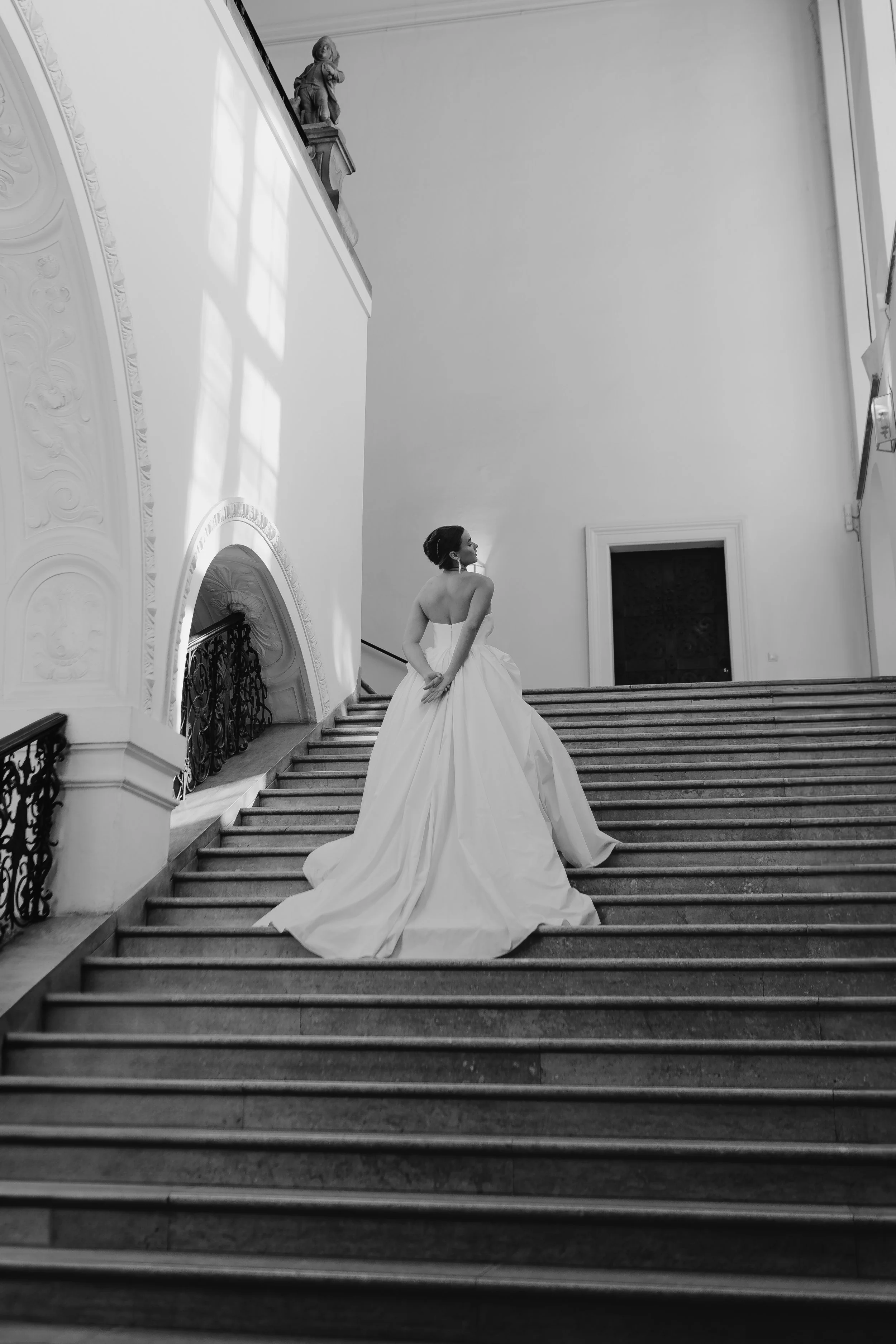 Eine junge Frau in einem eleganten weißen Brautkleid steht auf einer großen Treppe in einem prunkvollen, hell gestalteten Raum, der vermutlich in einem historischen Gebäude liegt.