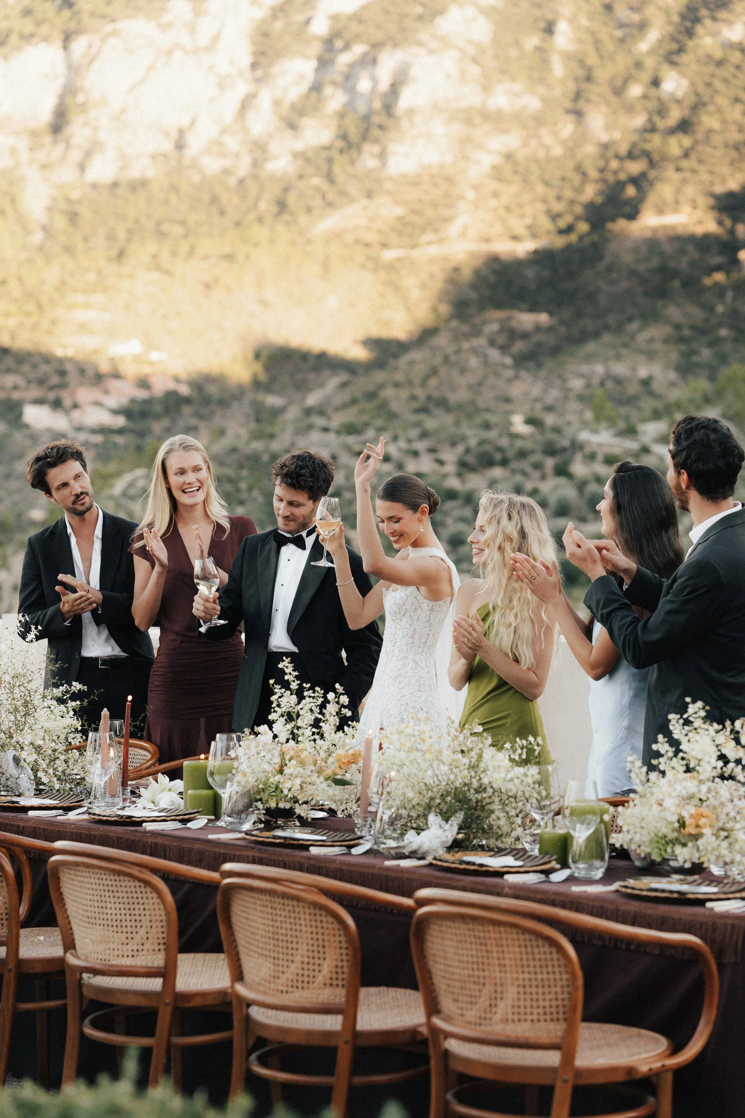 Menschen bei einer eleganten Feier am langen festlich gedeckten Tisch mit Blumen und Kerzen, feiern bei Sonnenuntergang, im Freien vor einer Bergkulisse.