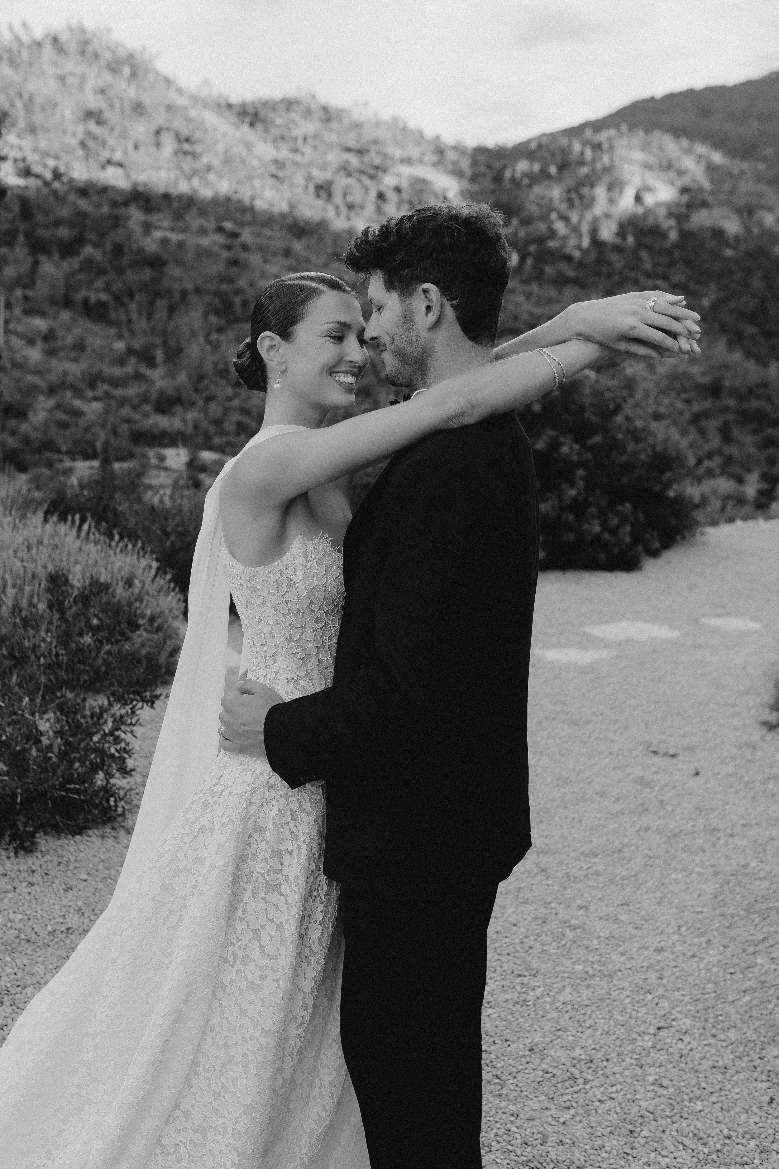 Ein Brautpaar umarmt sich in einer ländlichen Kulisse, beide lächeln entspannt.