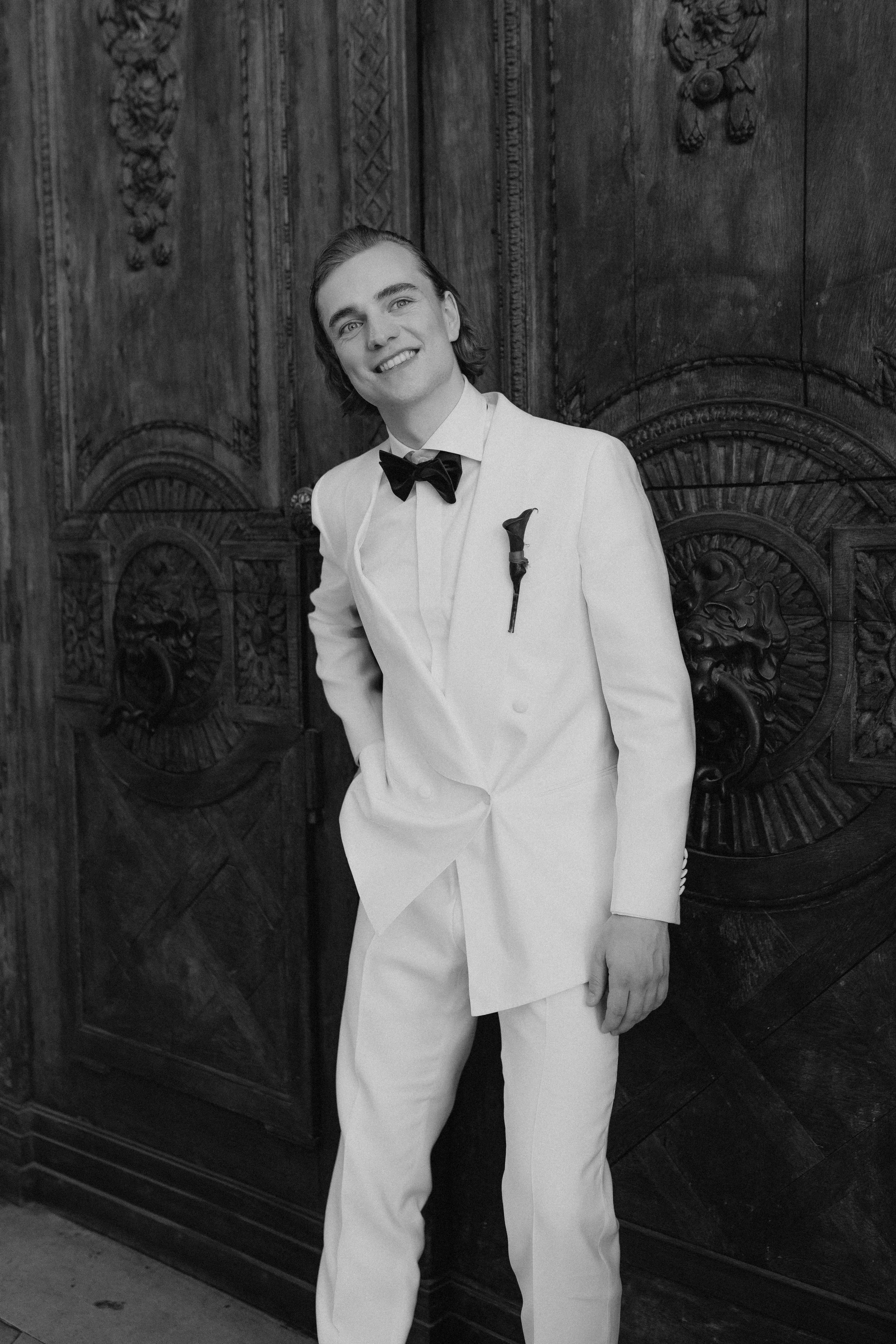 Ein junger Mann in einem weißen Anzug mit Fliege steht vor einem detaillierten, dunklen Holzportaltor, lächelt in die Kamera.