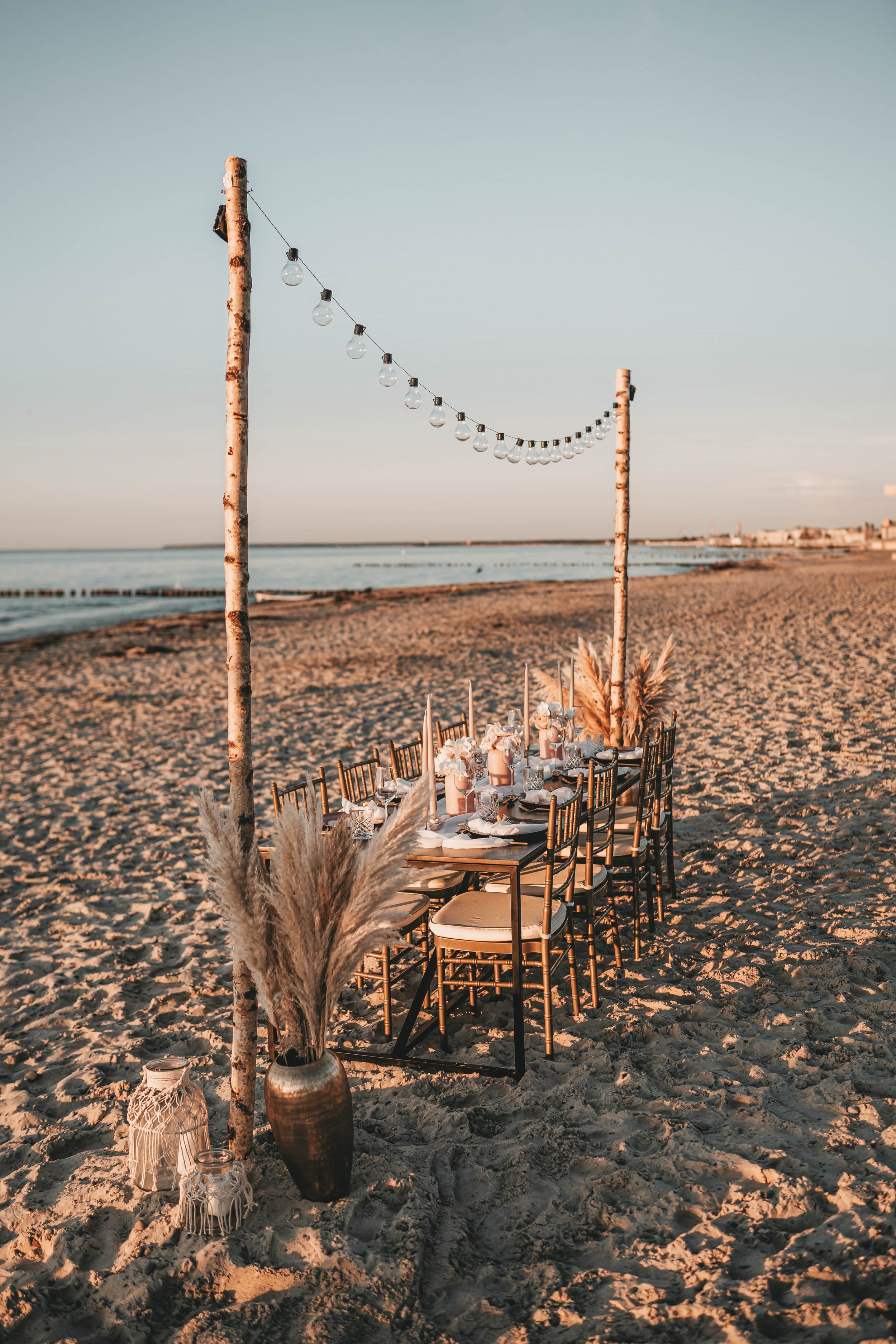 Elegantes, festlich gedecktes Tischset am Strand mit Lichtergirlande, pampasgras Dekoration und Sonnenuntergang.