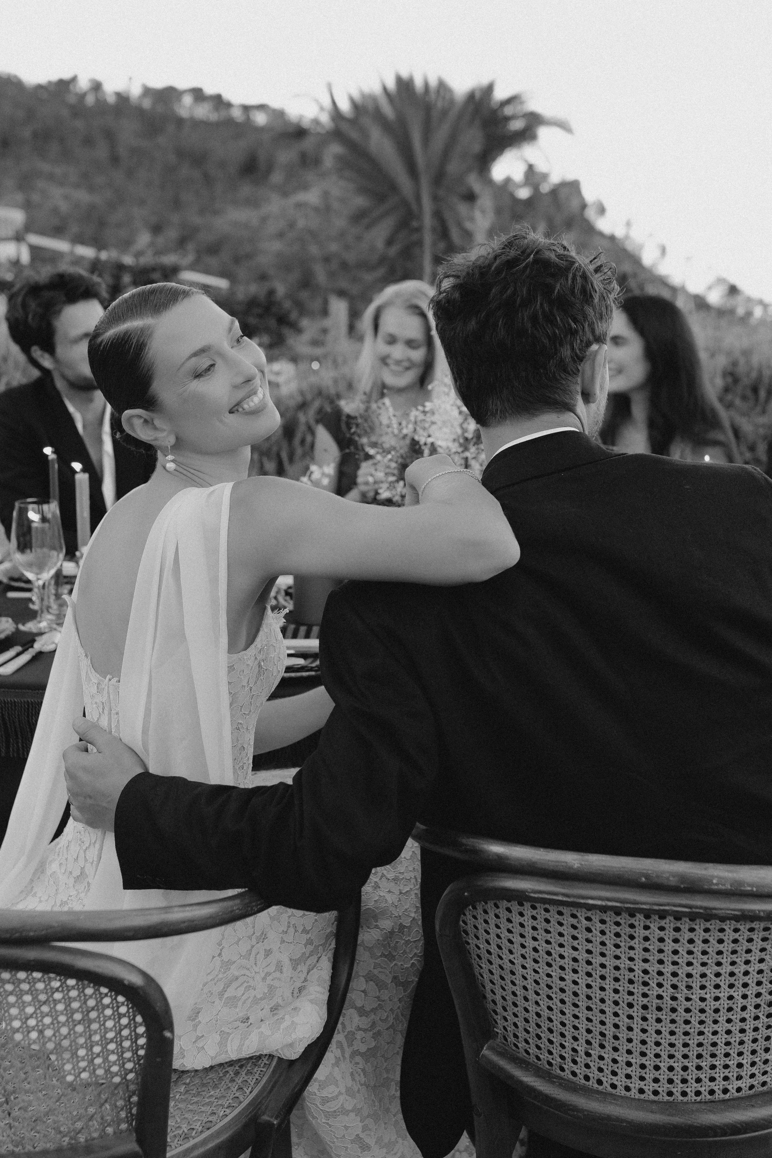 Eine Hochzeitsfeier im Freien, schwarze und weiße Fotografie, zartes Lächeln einer Braut, die auf einem Mann sitzt, umgeben von Freunden, bei Sonnenuntergang.