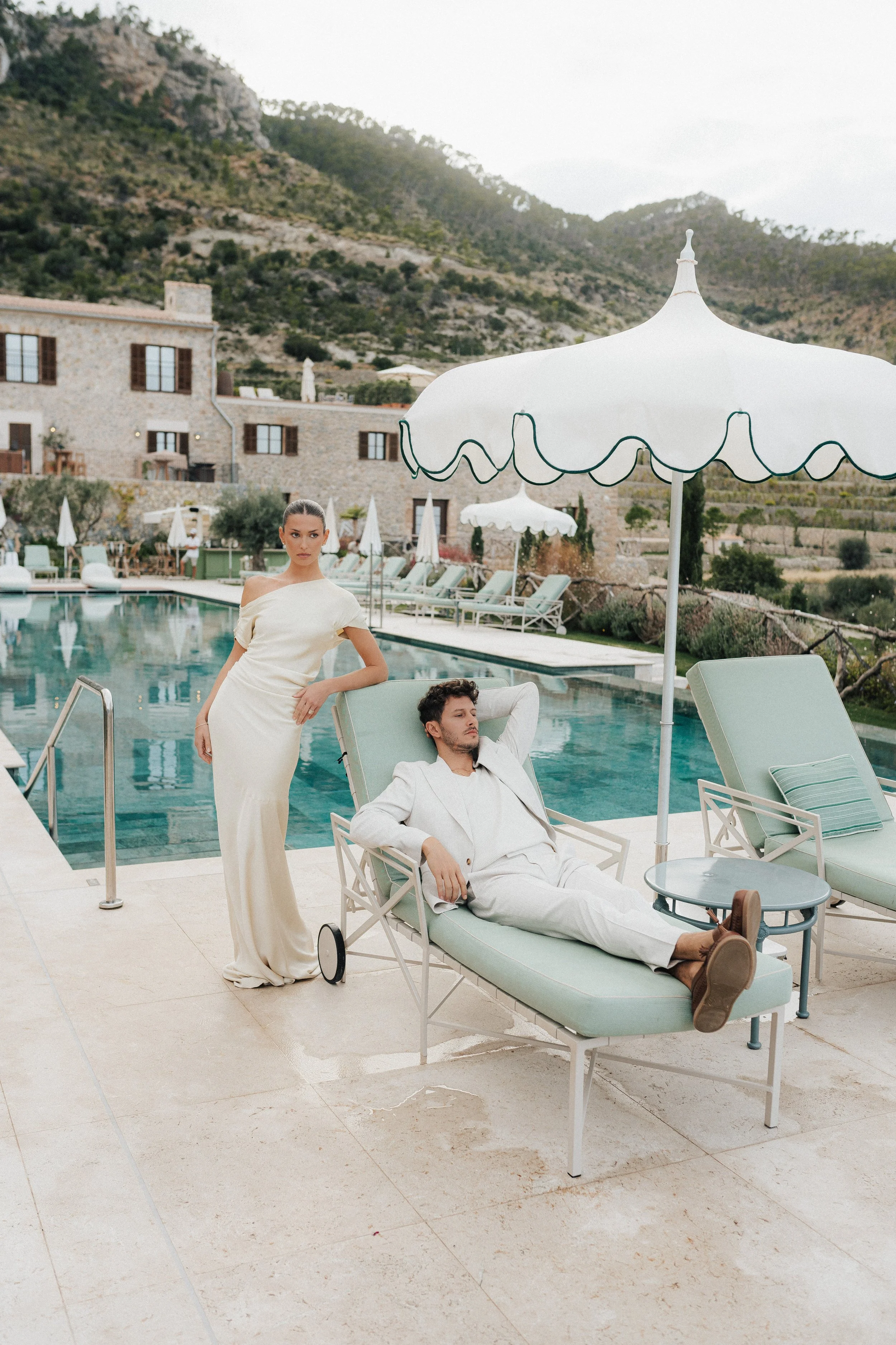 Ein Mann liegt entspannt auf einem Liegestuhl am Pool, während eine Frau in einem weißen Kleid neben ihm steht. Im Hintergrund sieht man Berge, ein Pool und Liegestühle mit Sonnenschirmen.