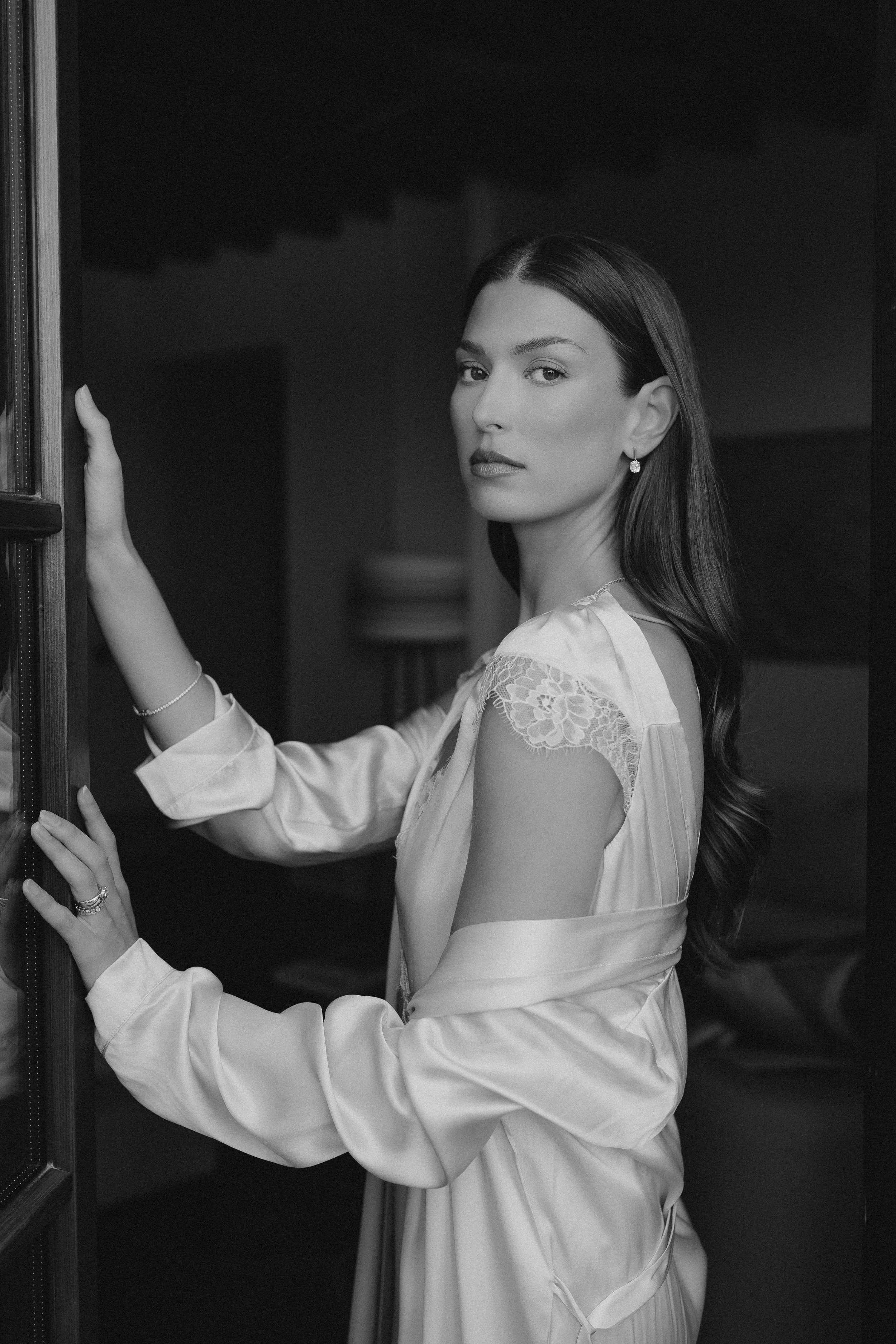 Schwarz-weiße Aufnahme einer jungen Frau mit langen, lockigen Haaren, die elegante Kleidung trägt und an einem Regal lehnt.