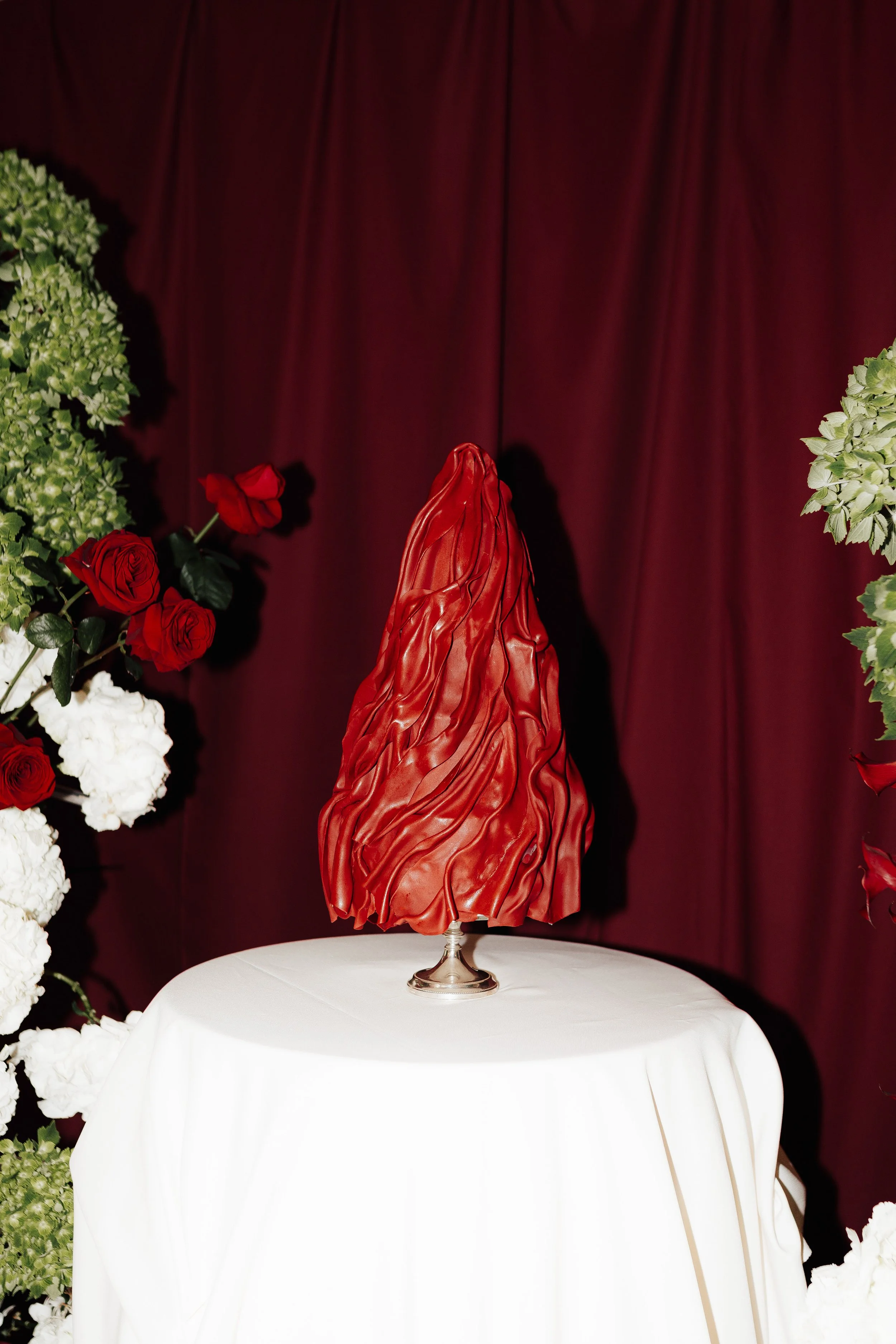 Eine kunstvolle Skulptur in roter Farbe auf einem weißen Tischtuch, umgeben von roten und weißen Blumen vor einem roten Vorhang.
