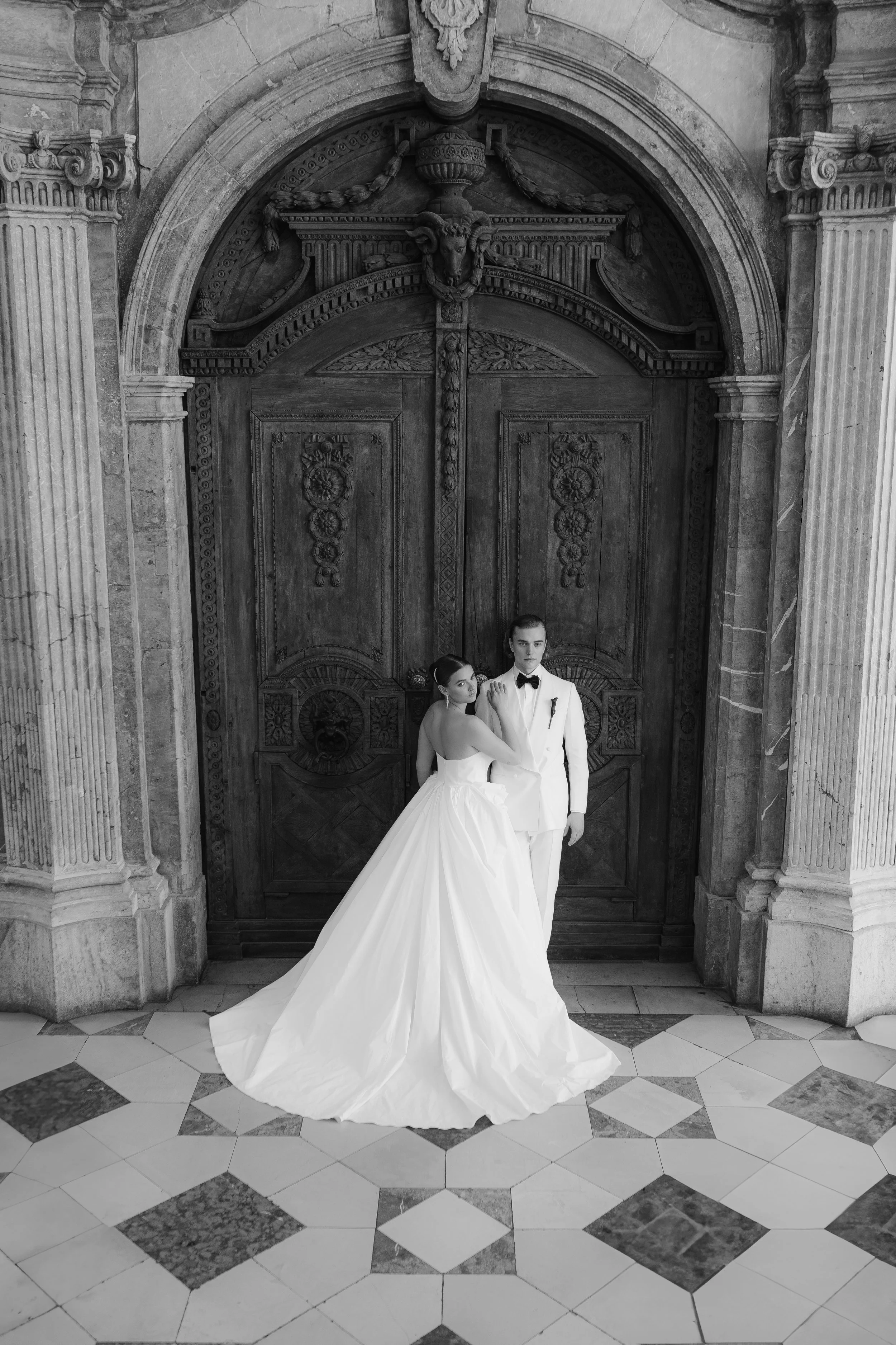 Braut und Bräutigam in formeller Kleidung vor einer großen, alten Holztür in einem historischen Gebäude, schwarz-weiß Fotografie.