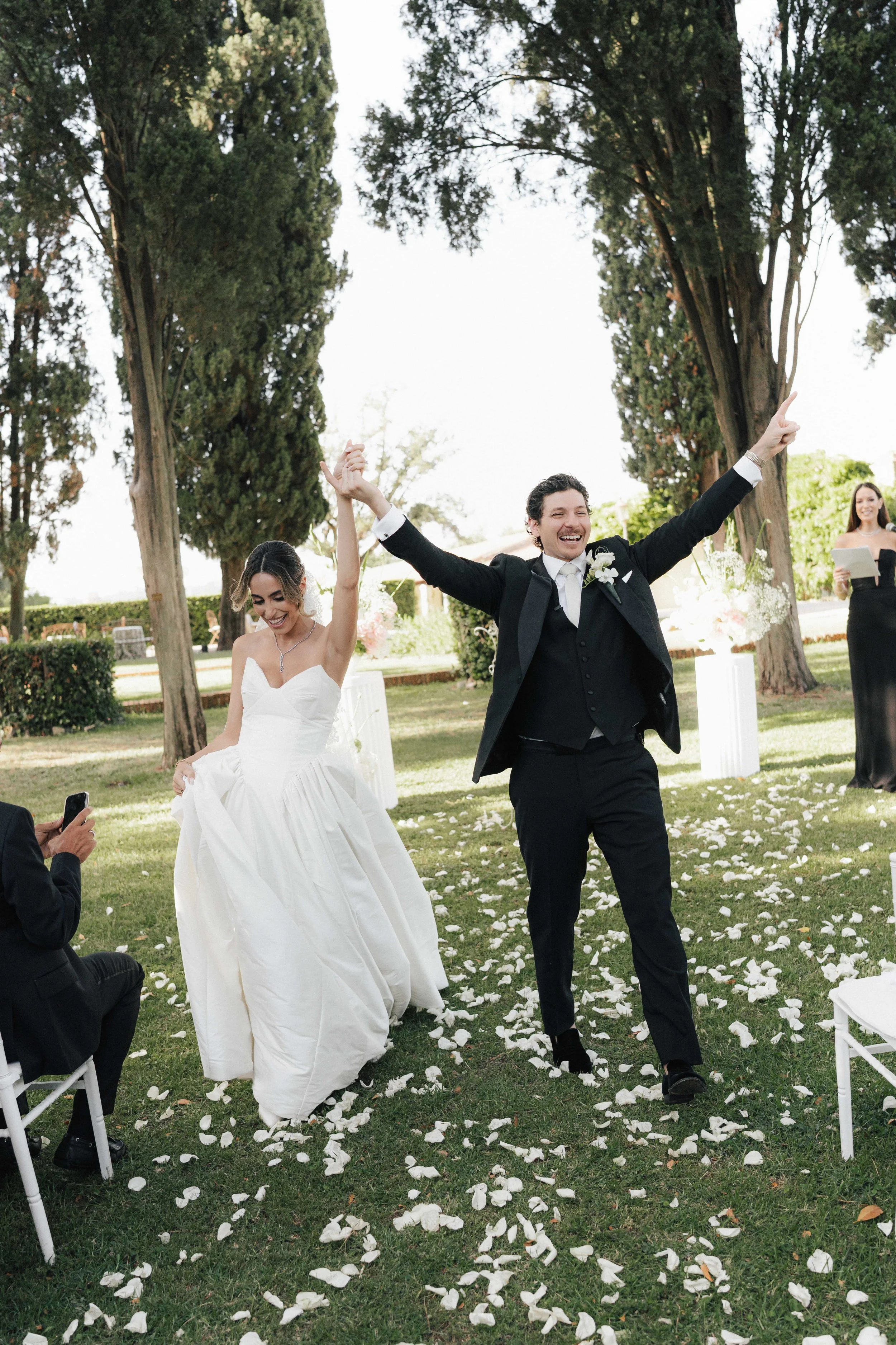 Ein lachendes Brautpaar beim Applaus während ihrer Hochzeit im Freien, mit weißen Blütenblättern auf dem Rasen.