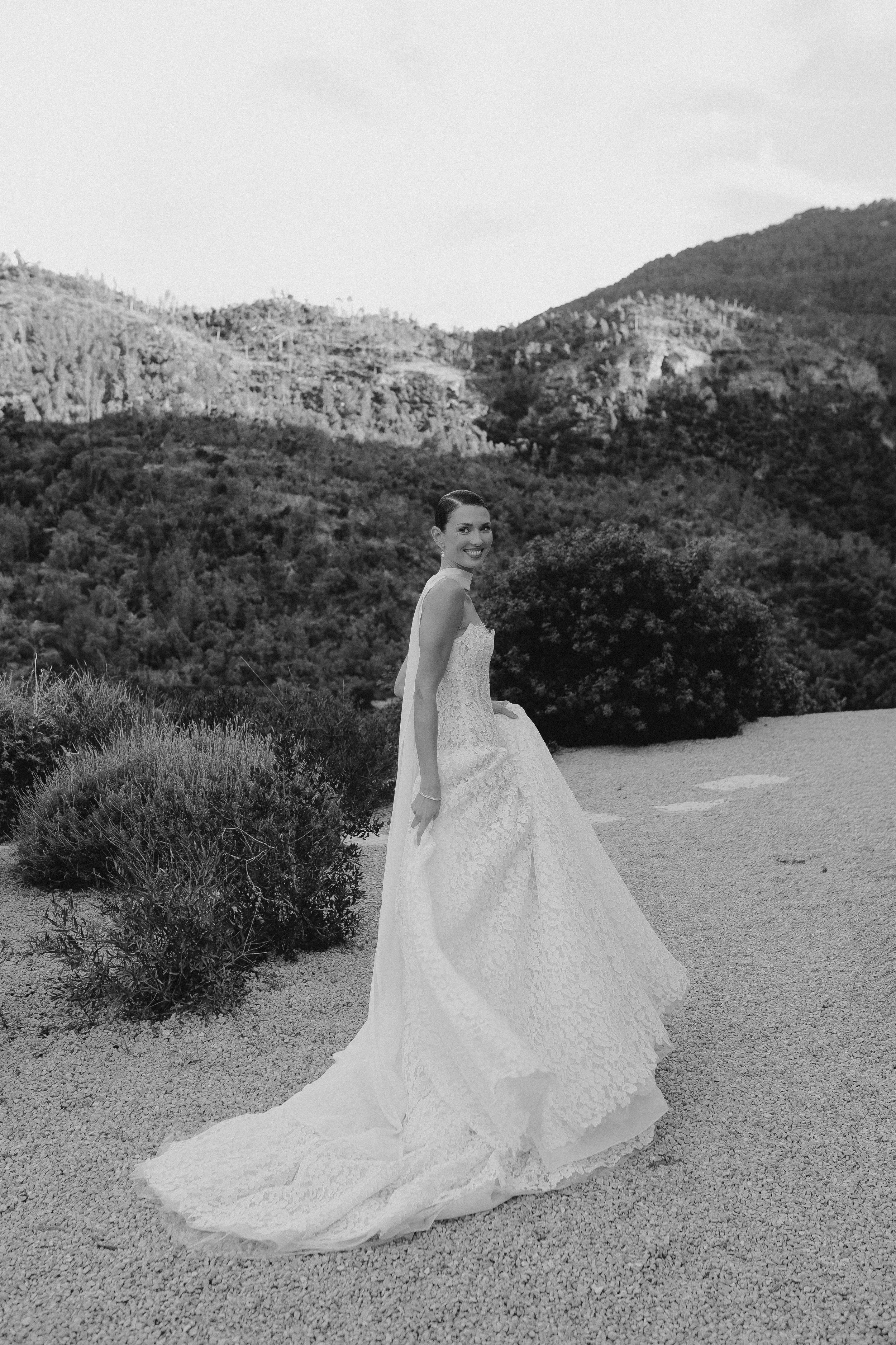 Eine Frau in einem Hochzeitskleid steht in einer natürlichen, bergigen Landschaft mit Büschen und Hügeln im Hintergrund.