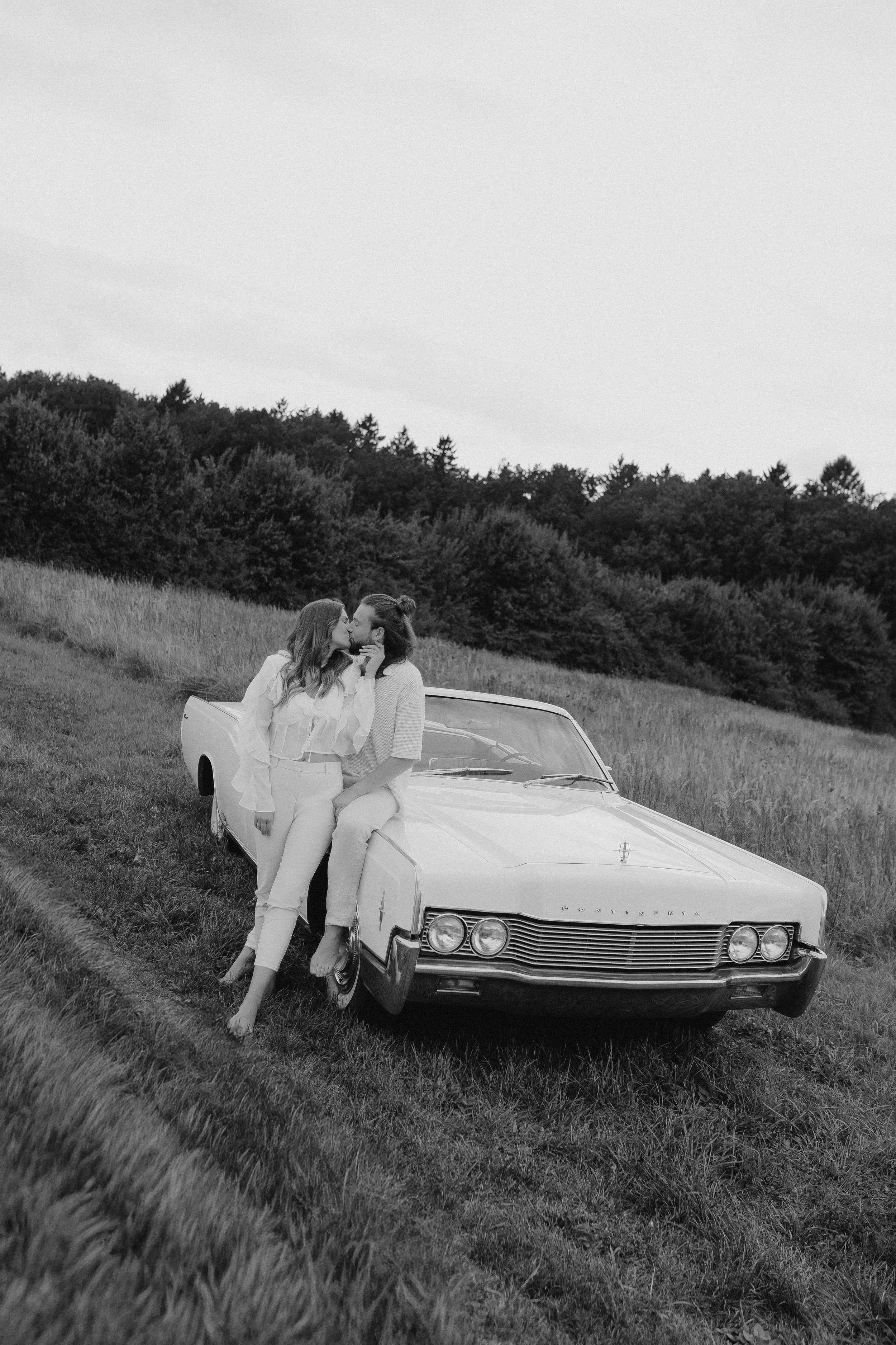 Zwei Frauen küssen sich an einem alten Auto in einer offenen Wiese, umgeben von Bäumen, in Schwarz-Weiß.