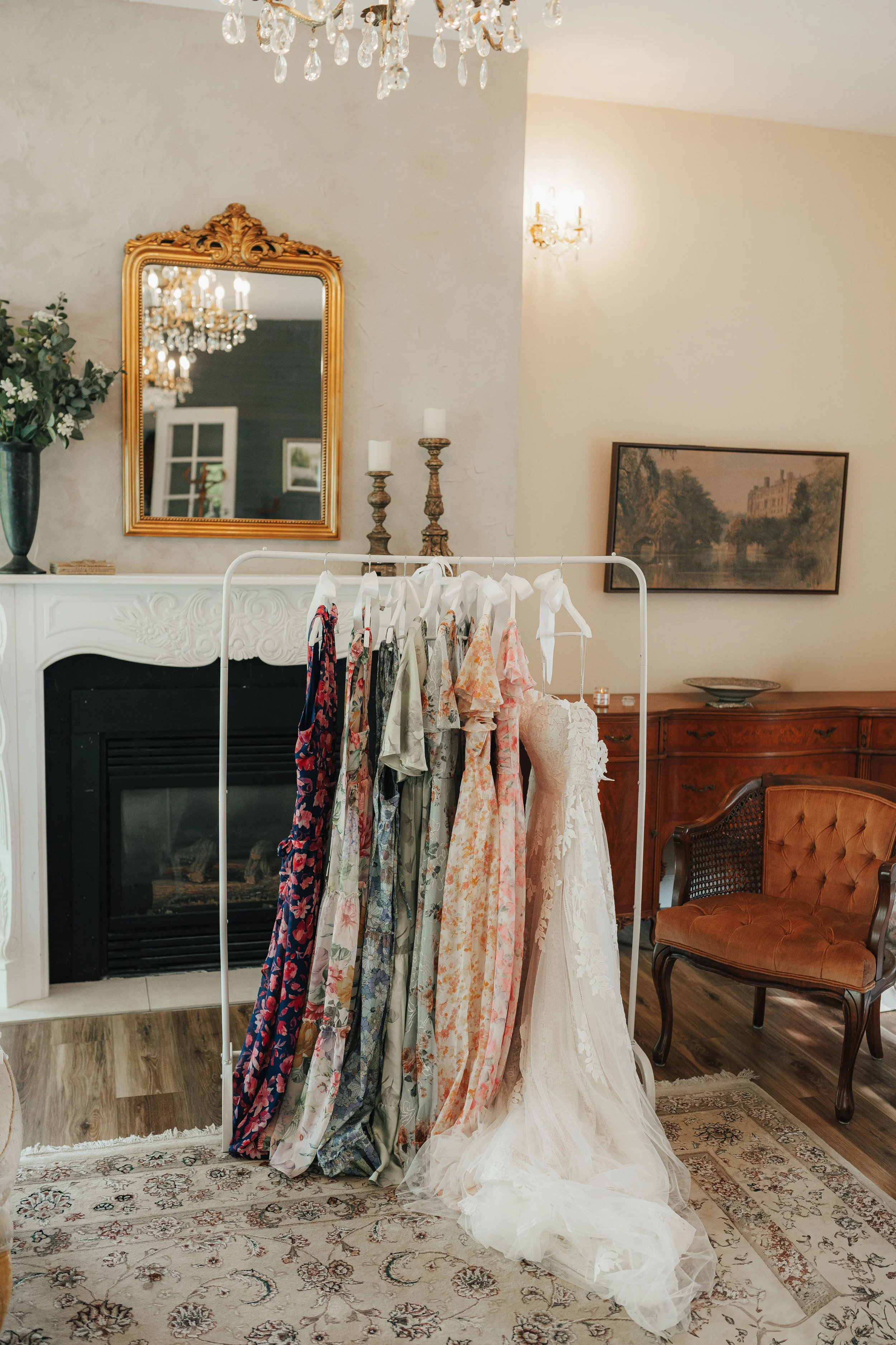 Hochzeitskleider und Sommerkleider hängen an einer Kleiderstange vor einem offenen Kamin in einem eleganten, gut dekorierten Wohnzimmer.