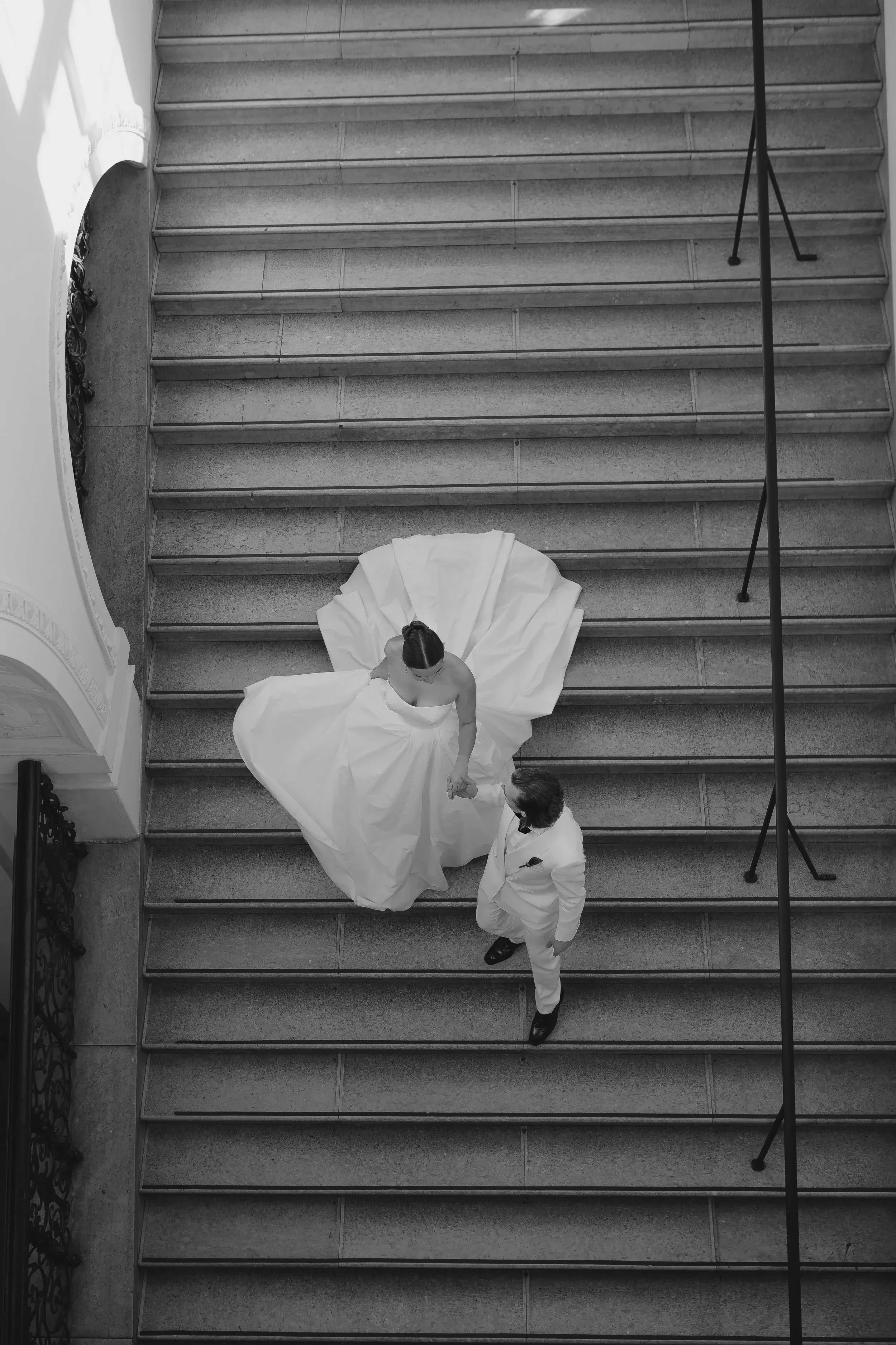 Braut in weißem Brautkleid auf einer Treppe mit einem Mann in weißem Anzug, aus einer Vogelperspektive fotografiert.