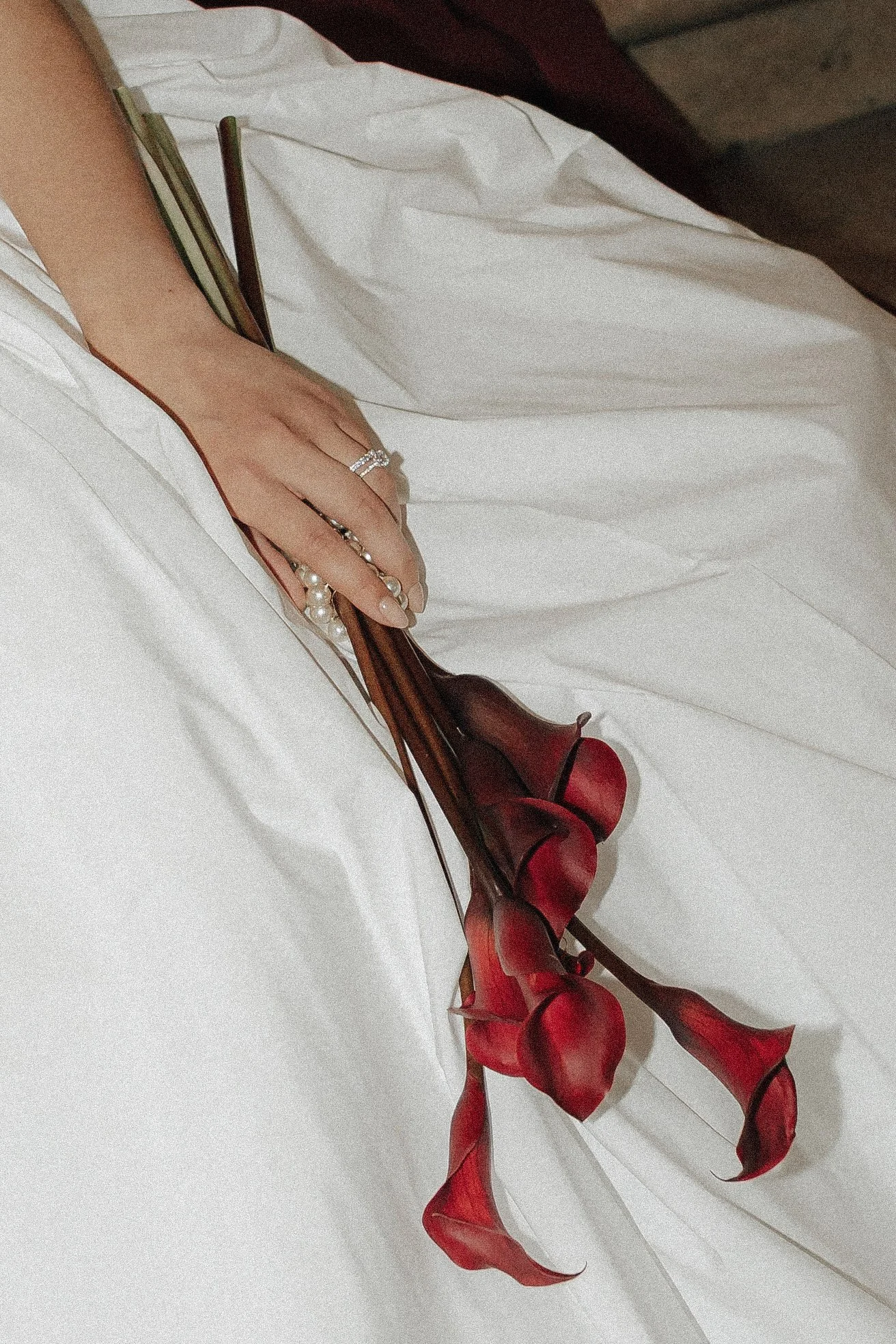 Hand mit Ehering hält rote Calla-Lilien auf weißem Stoff.