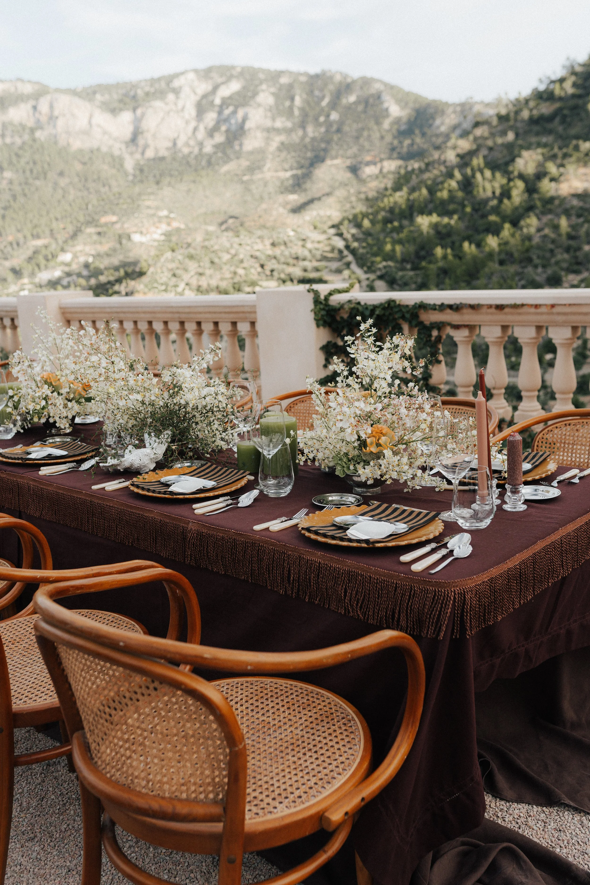 Ein festlich gedeckter Tisch mit Blumen, Kerzen und Geschirr vor einer Bergkulisse auf einer Terrasse.