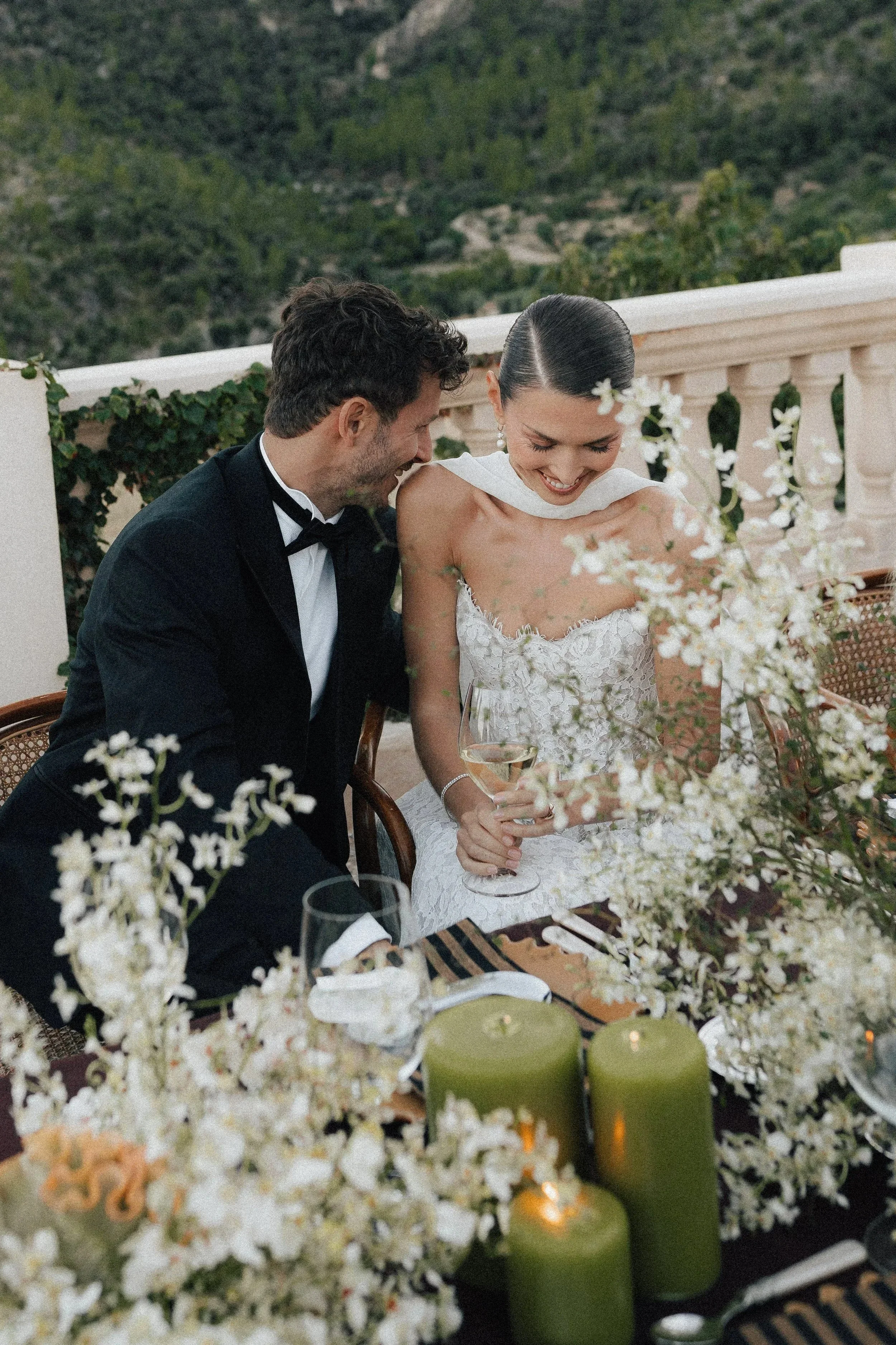 Ein glückliches Brautpaar bei ihrer Hochzeit, sitzend an einem festlich gedeckten Tisch mit weißen Blumen und grünen Kerzen, im Freien mit Berglandschaft im Hintergrund.