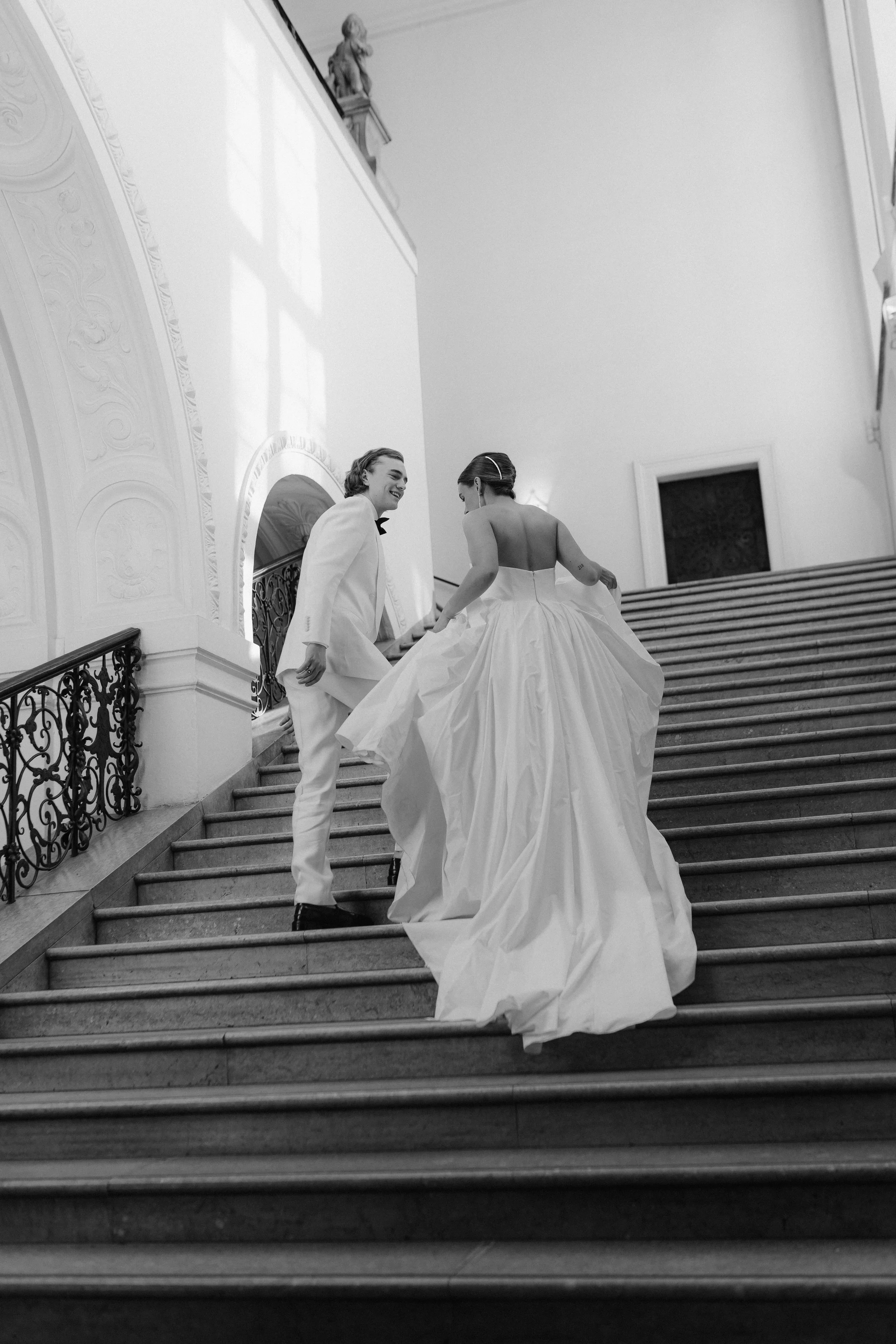 Zwei Personen in formeller Kleidung, eine Frau in Brautkleid und ein Mann in Anzug, lachen und steigen die Treppe in einem eleganten, historisch wirkenden Gebäude.