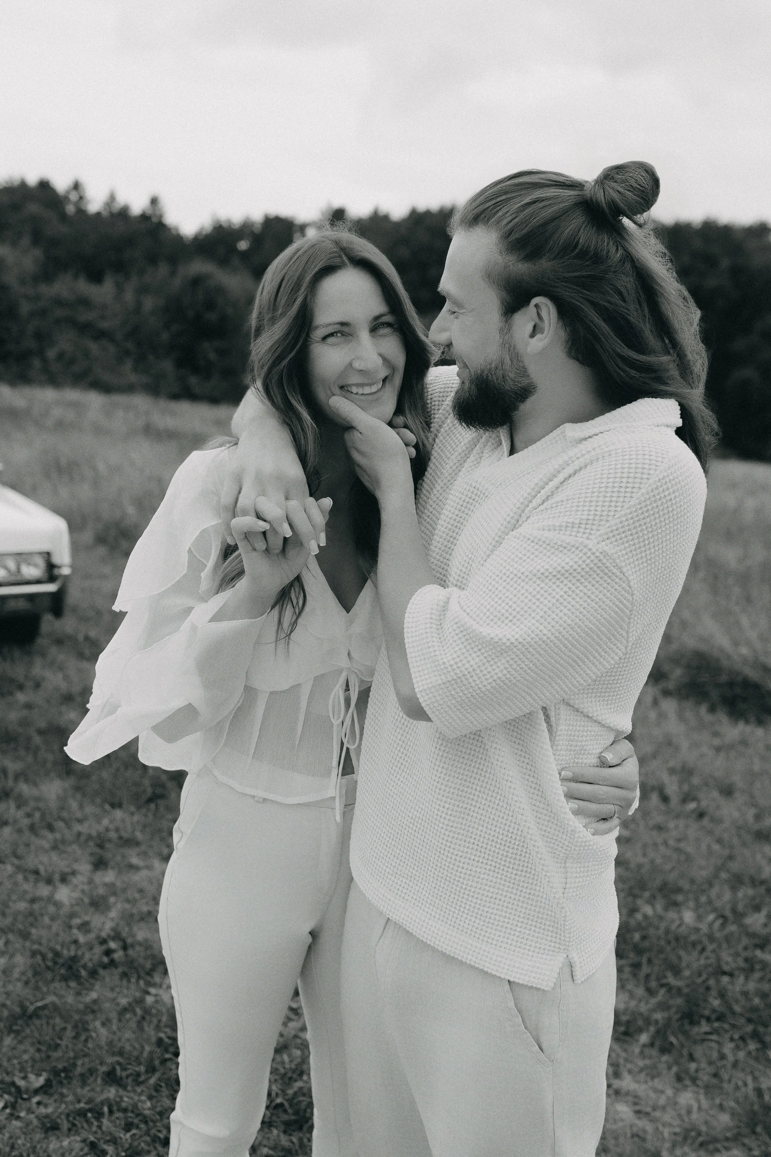Ein glückliches Paar umarmt sich im Freien auf einer Wiese, lächelt und schaut sich liebevoll an, im Hintergrund Bäume und Wolken.