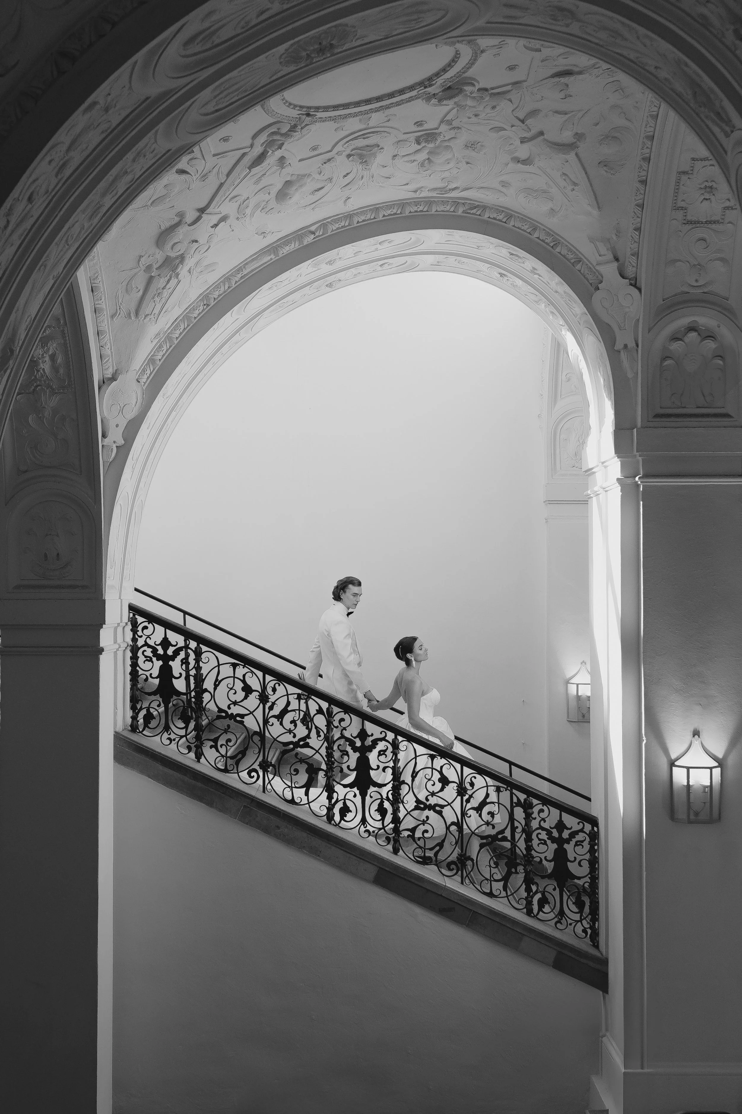 Ein Brautpaar auf einer verzweigten, stilvollen Treppe in einem prunkvollen, historischen Gebäude, schwarz-weiß Fotografie.