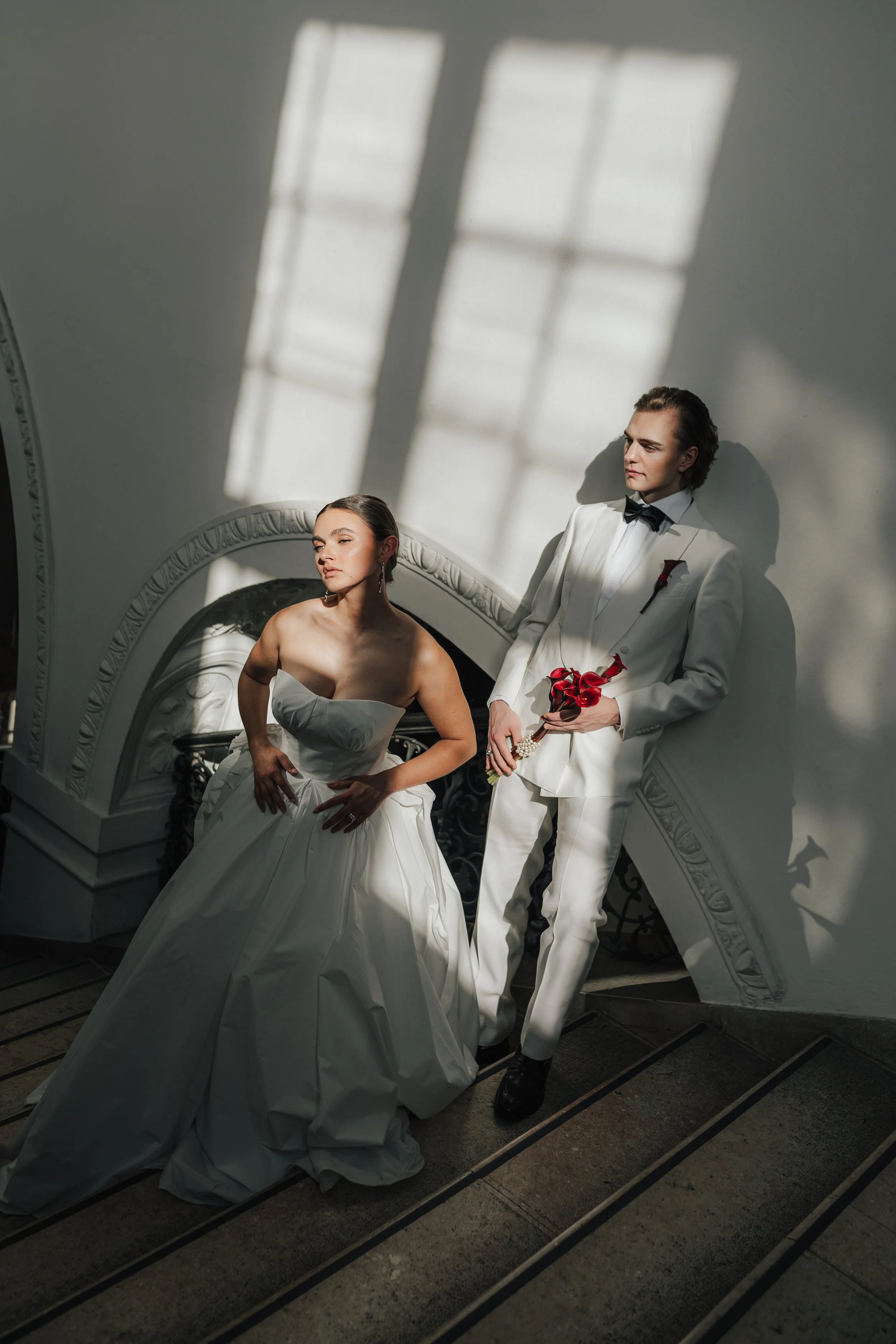 Ein Brautpaar in formeller Kleidung, die Frau in einem weißen Brautkleid und der Mann in einem weißen Anzug mit Fliege, stehen auf einer Treppe im Sonnenlicht, das durch ein Fenster fällt.
