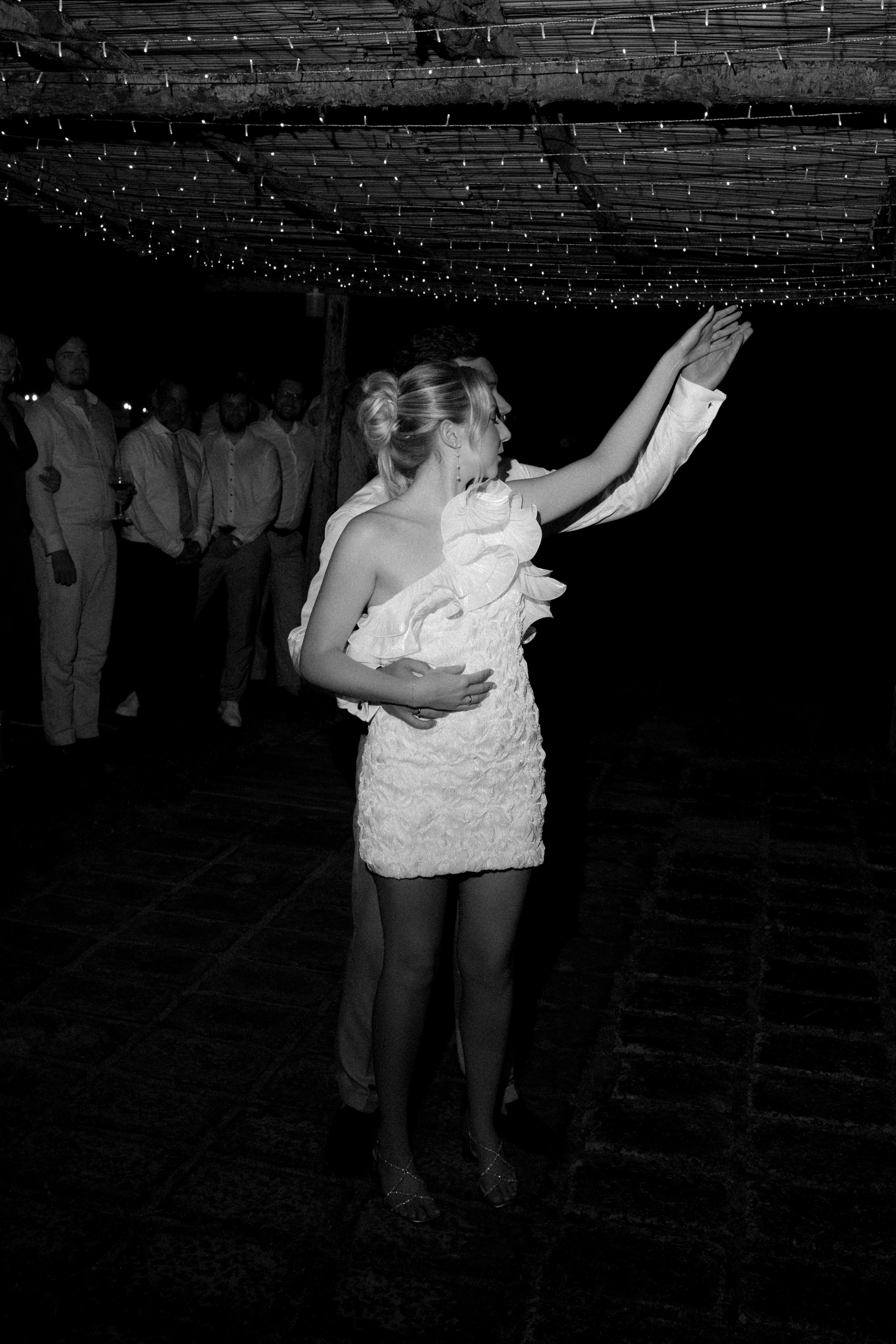 Eine Braut in einem Spitzenkleid tanzt, umgeben von Gästen bei einer Hochzeit im Freien bei Nacht, beleuchtet von Lichterketten unter einer hölzernen Überdachung.