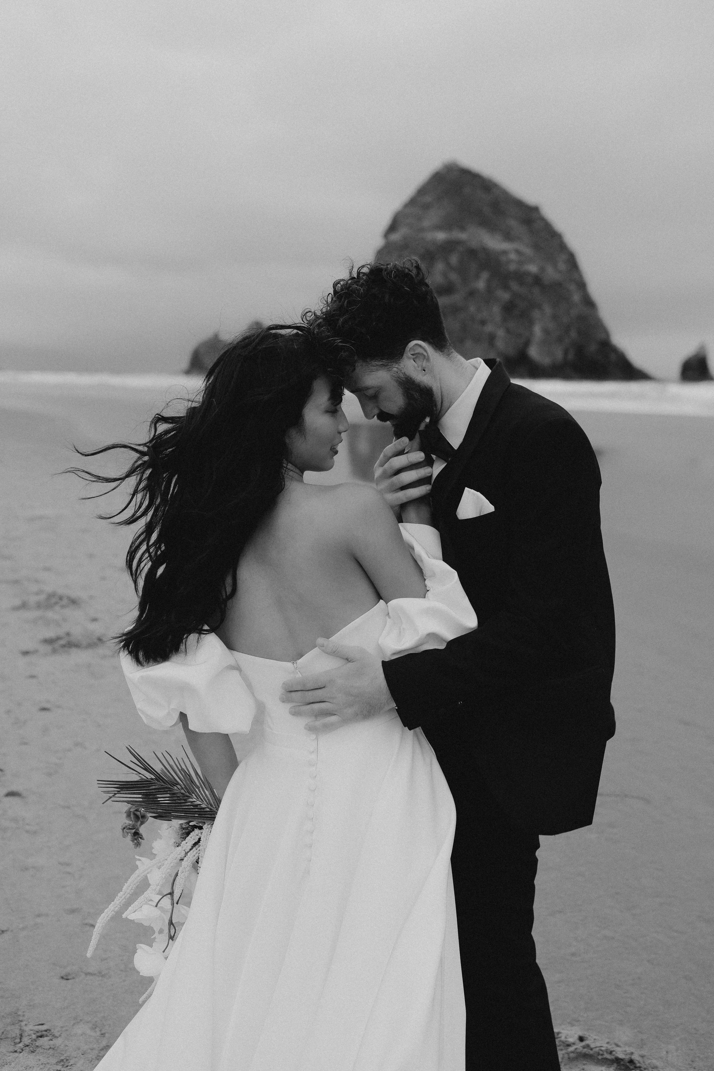 Ein glückliches Paar in formeller Kleidung am Strand bei Sonnenuntergang, umgeben von Felsen im Hintergrund.