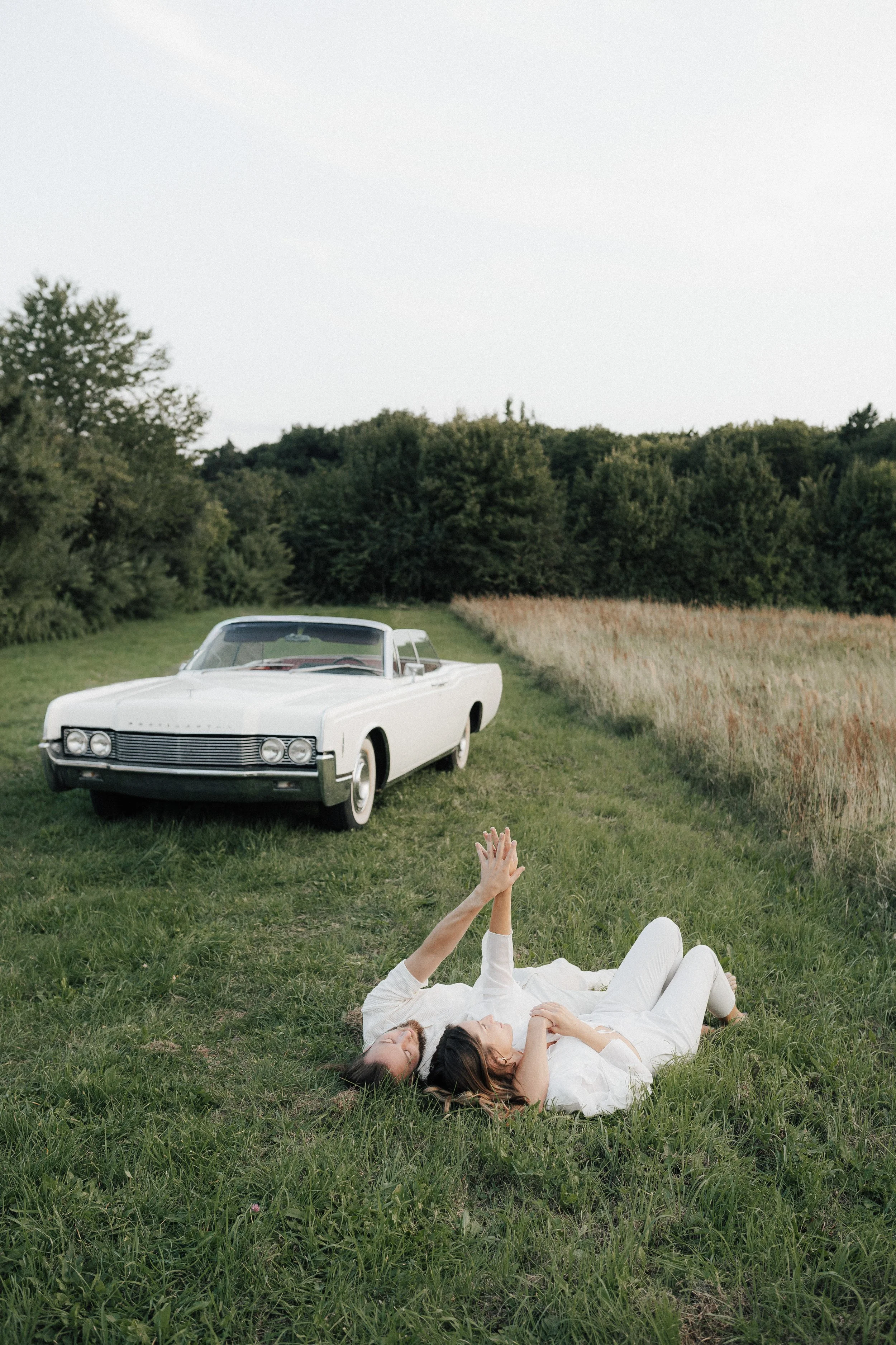 Zwei Frauen liegen entspannt auf dem Gras in einem offenen Feld, während sich im Hintergrund ein weißer Oldtimer-Cabriolet befindet.