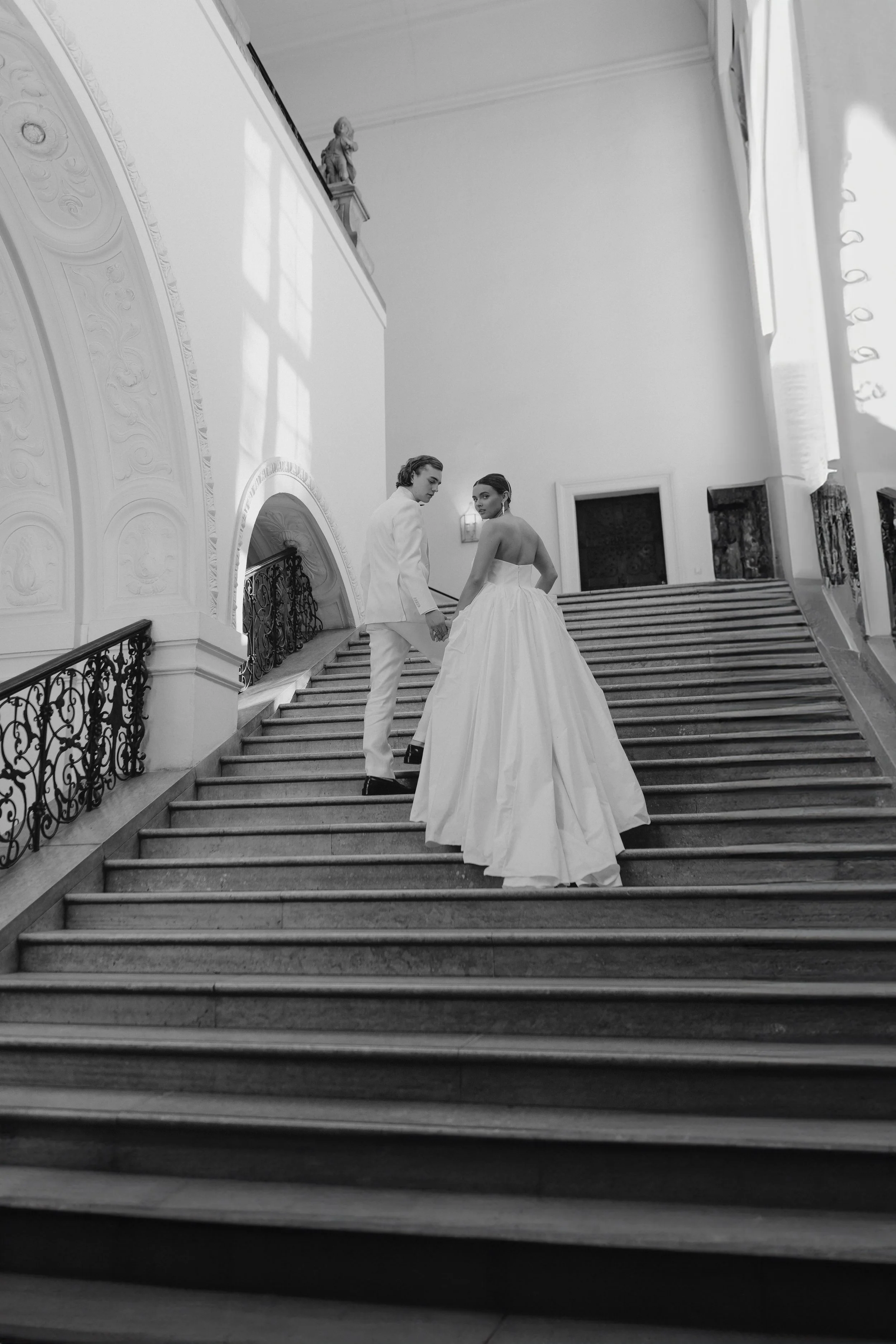 Braut und Bräutigam auf einer prächtigen Treppe in einem eleganten Gebäude, beide in Hochzeitskleidung, Hand in Hand zeigend auf die Kamera.