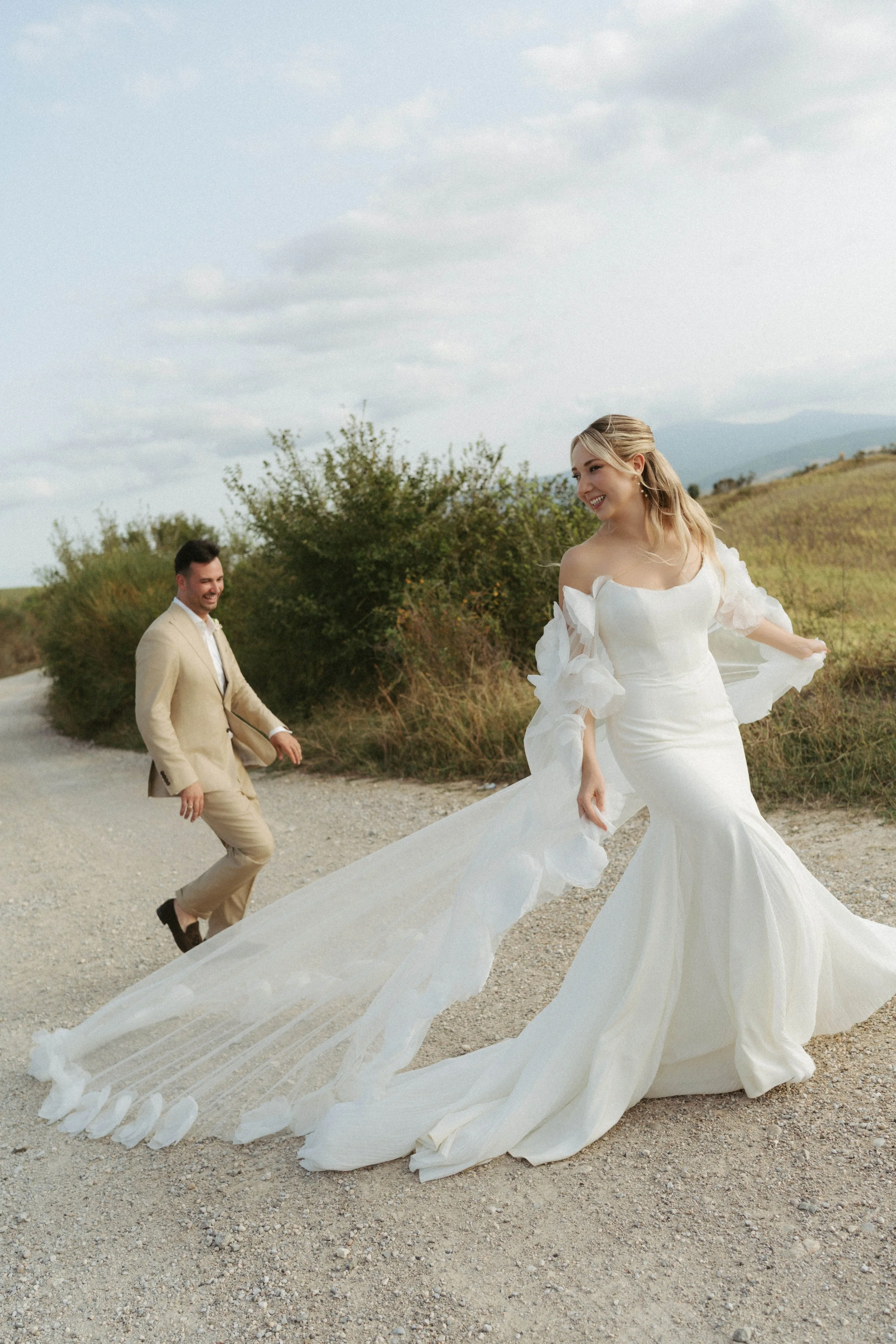 Ein glückliches Paar bei einer Hochzeitsfeier in einer ländlichen Gegend mit grünem Hügel im Hintergrund. Die Frau trägt ein elegantes, langes weißes Hochzeitskleid, während der Mann in einem beige Anzug gekleidet ist. Sie lachen und tanzen auf einem