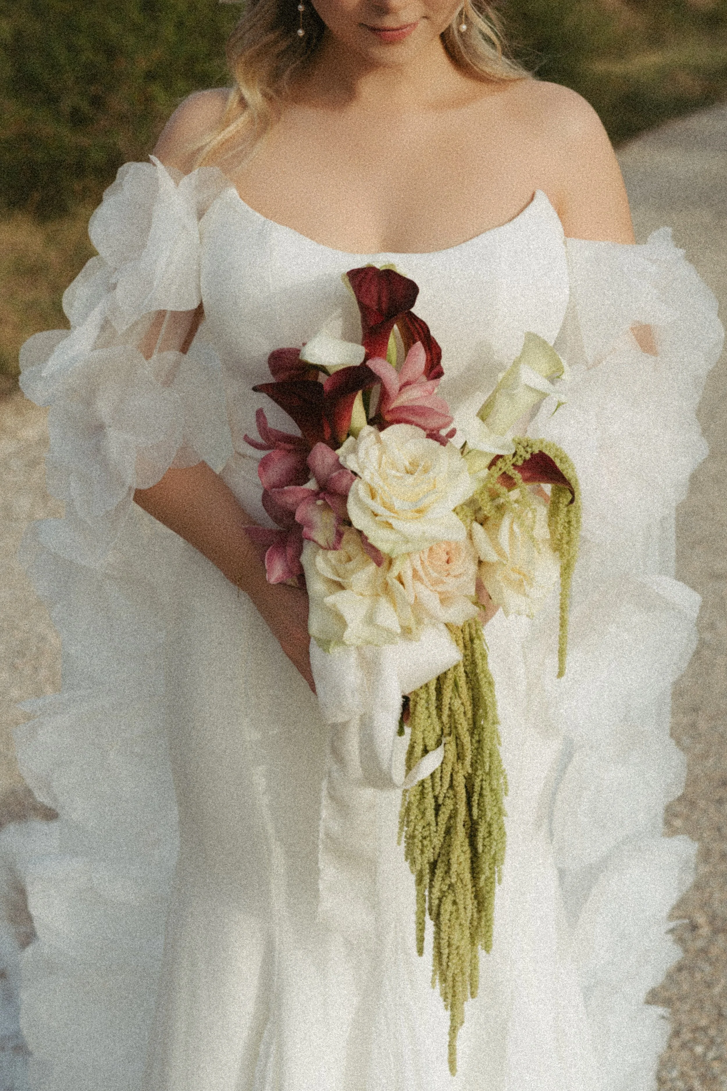 Frau in weißem Brautkleid hält einen Brautstrauß, bestehend aus weißen, roten, rosa und grünen Blumen, vor einem natürlichen Hintergrund.