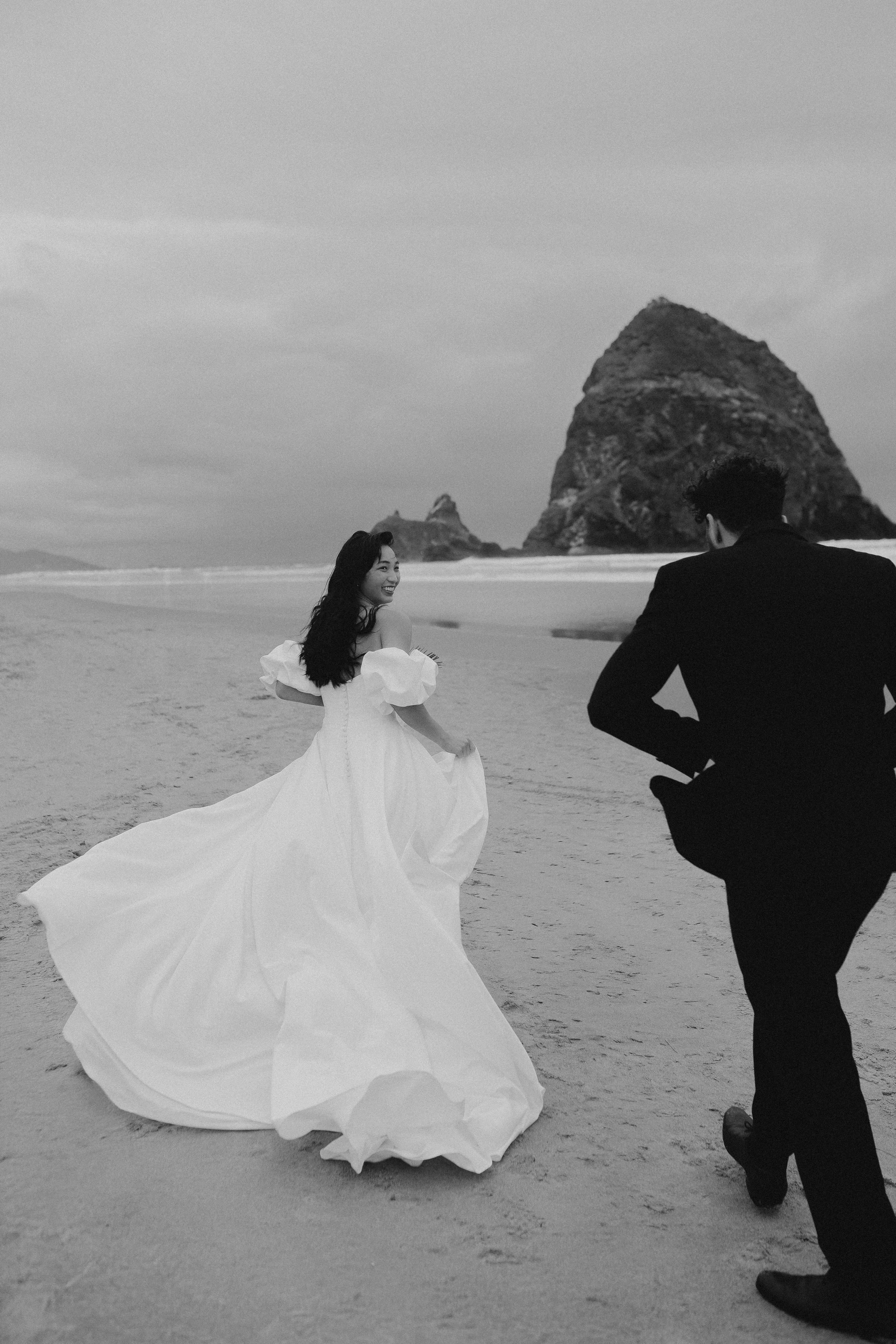 Paare in Hochzeitskleidung laufen am Strand entlang, mit Felsen im Hintergrund, in Schwarz-Weiß-Aufnahme.