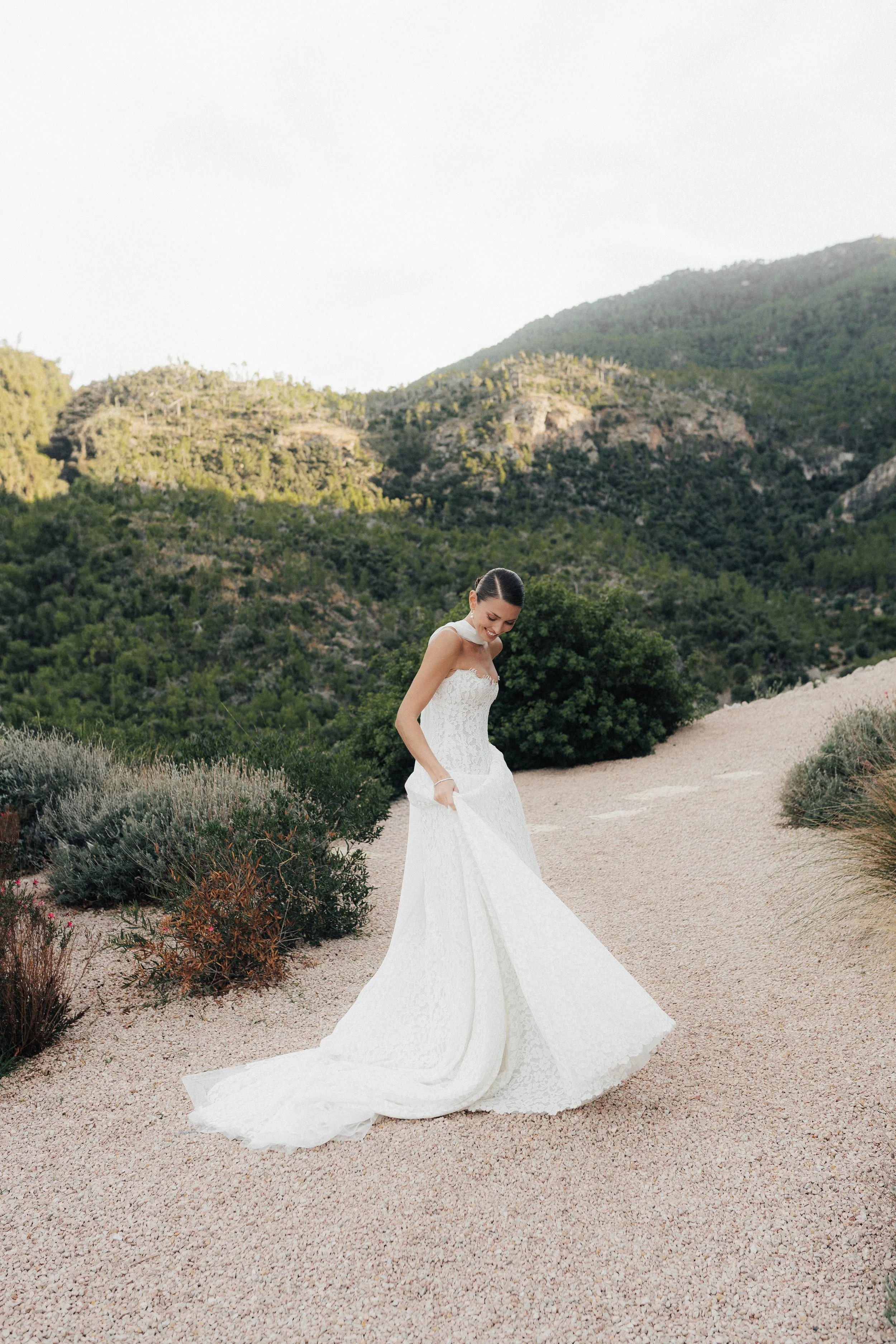Braut in Hochzeitskleid auf Weg in Natur mit Bergen im Hintergrund.