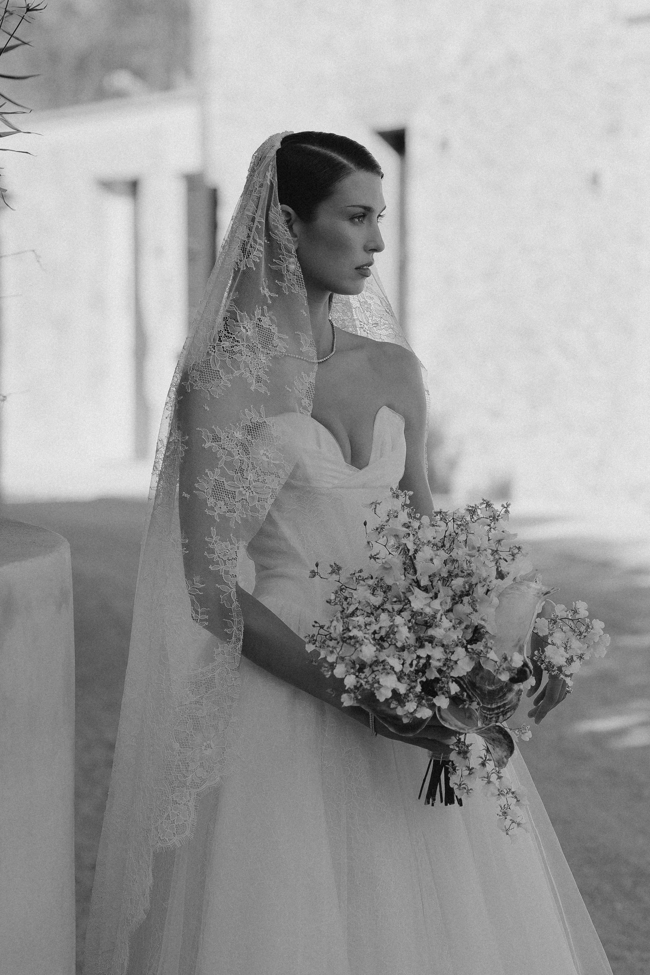 Schwarz-Weiß-Foto einer Frau in Hochzeitskleid mit Blumengebinde, stehend im Freien, mit Blick zur Seite.