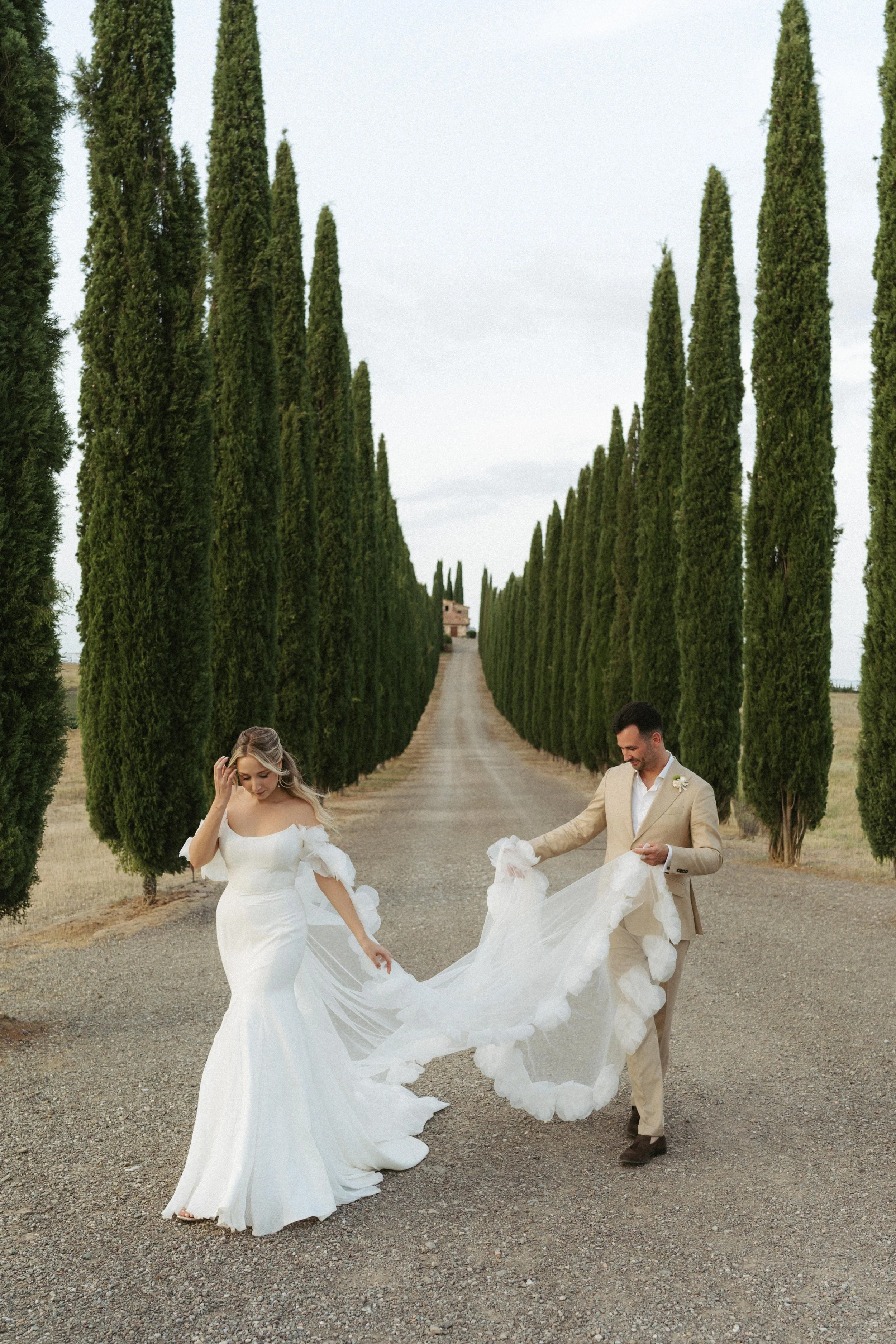 Braut und Bräutigam tanzen an einem Weg, umgeben von hohen, grünen Zypressen, bei einer Hochzeit im Freien.