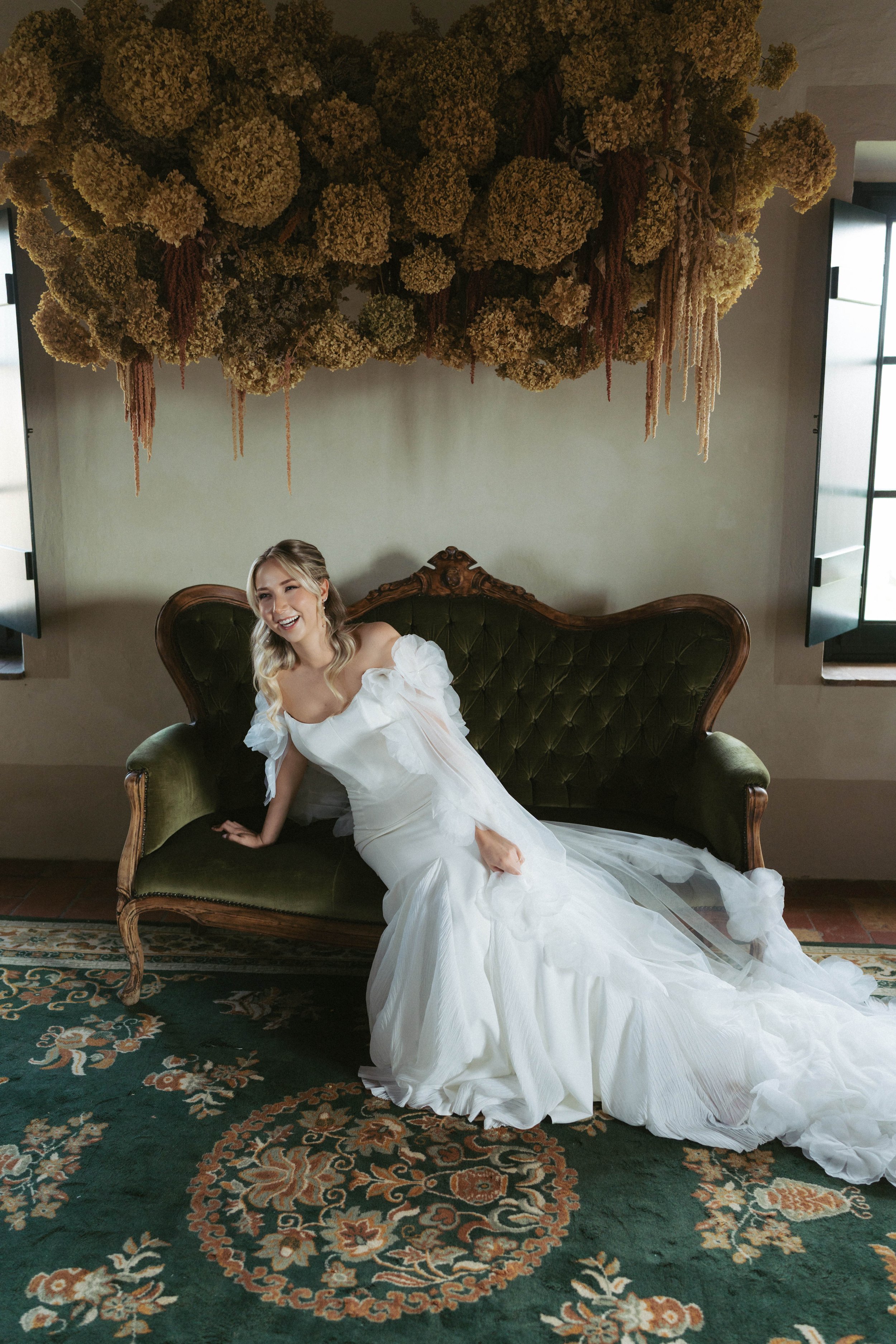 Eine lachende Frau in einem weißen Brautkleid, die auf einer antiken grünen Couch sitzt, mit großen getrockneten Blumen über ihr an der Decke