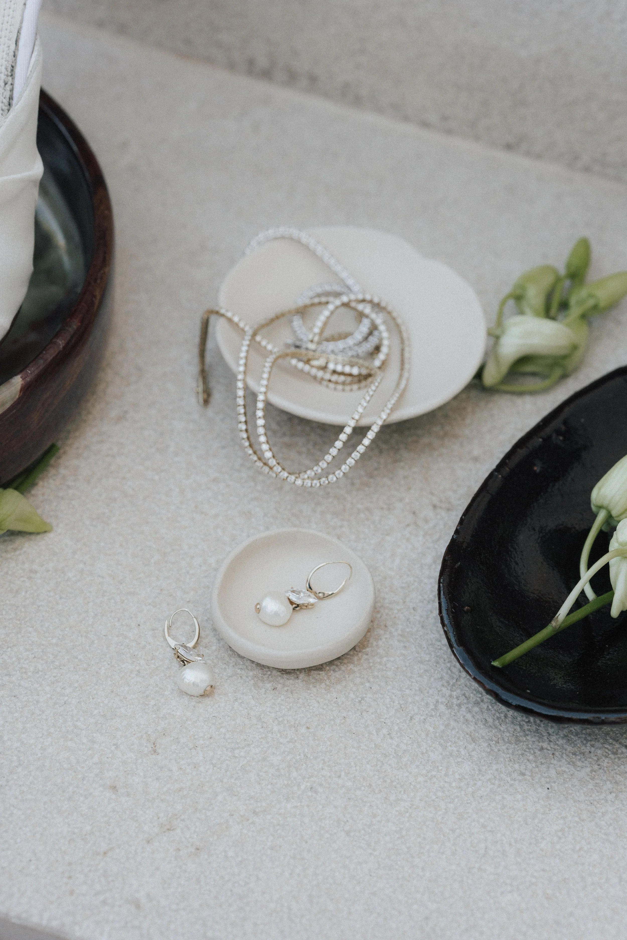 Schmuck, darunter Perlenohrringe und Halsketten, auf einer hellen Oberfläche, umgeben von Blumen und schwarzen Schalen.