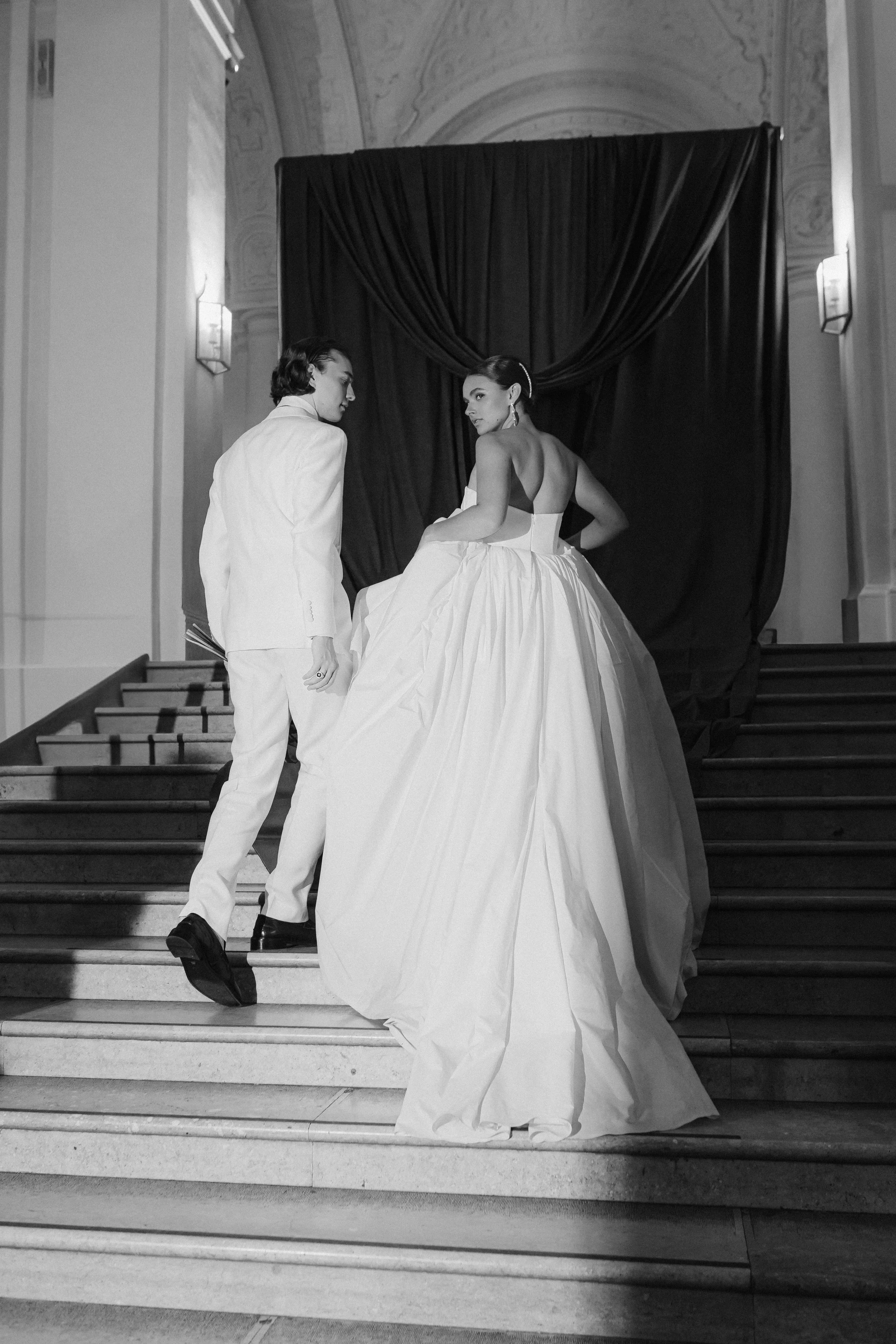 Schwarz-weiße Aufnahme eines Brautpaares auf einer Treppe, die Frau trägt ein voluminöses Brautkleid, der Mann einen weißen Anzug, im Hintergrund ein dunkler Vorhang.