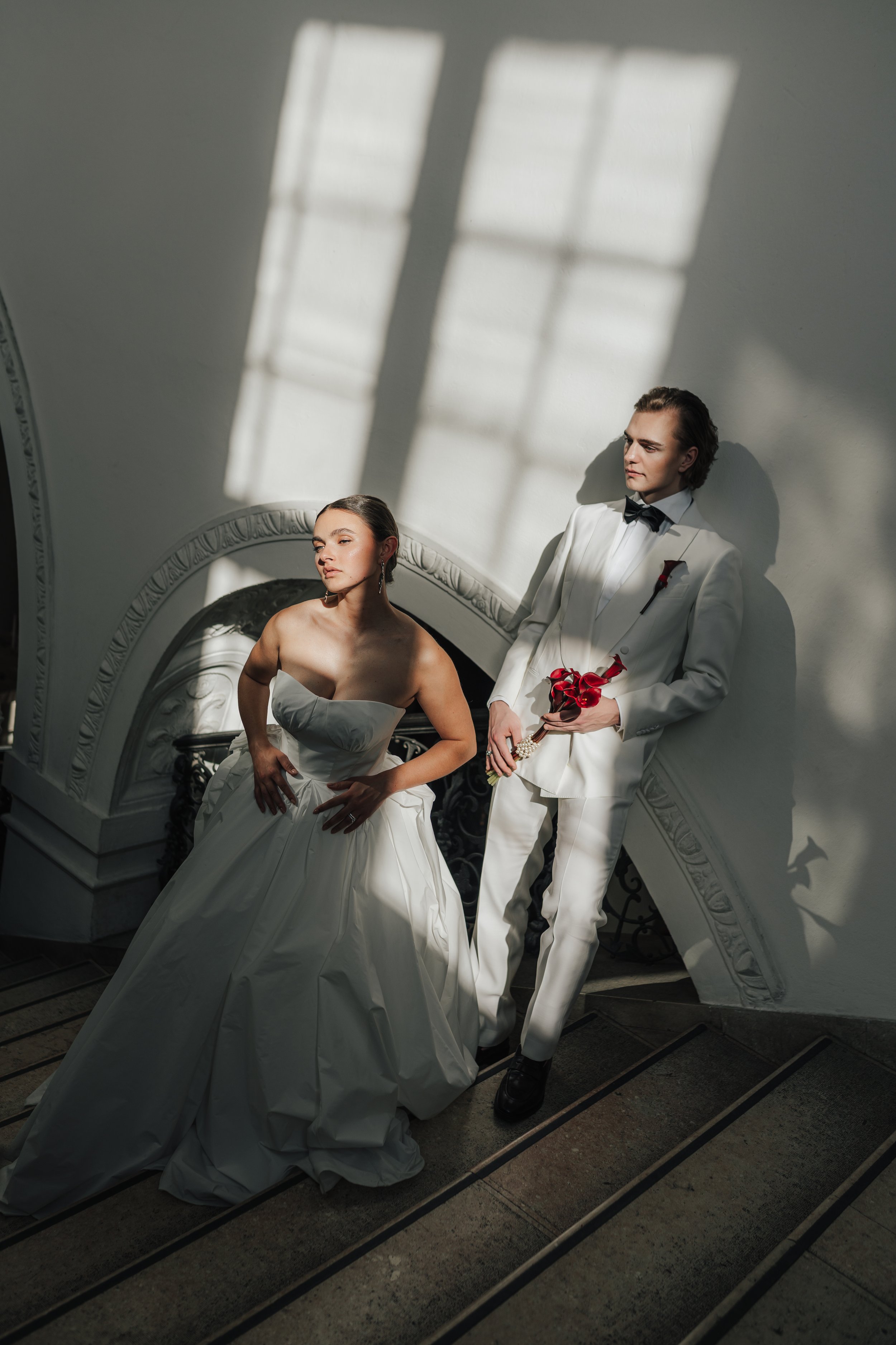 Ein junges Paar in Hochzeitskleidung steht in einem eleganten Treppenhaus. Die Frau trägt ein weißes Brautkleid und der Mann einen weißen Anzug mit Fliege. Sonnenlicht fällt durch ein Fenster und erzeugt Schatten auf der Wand.