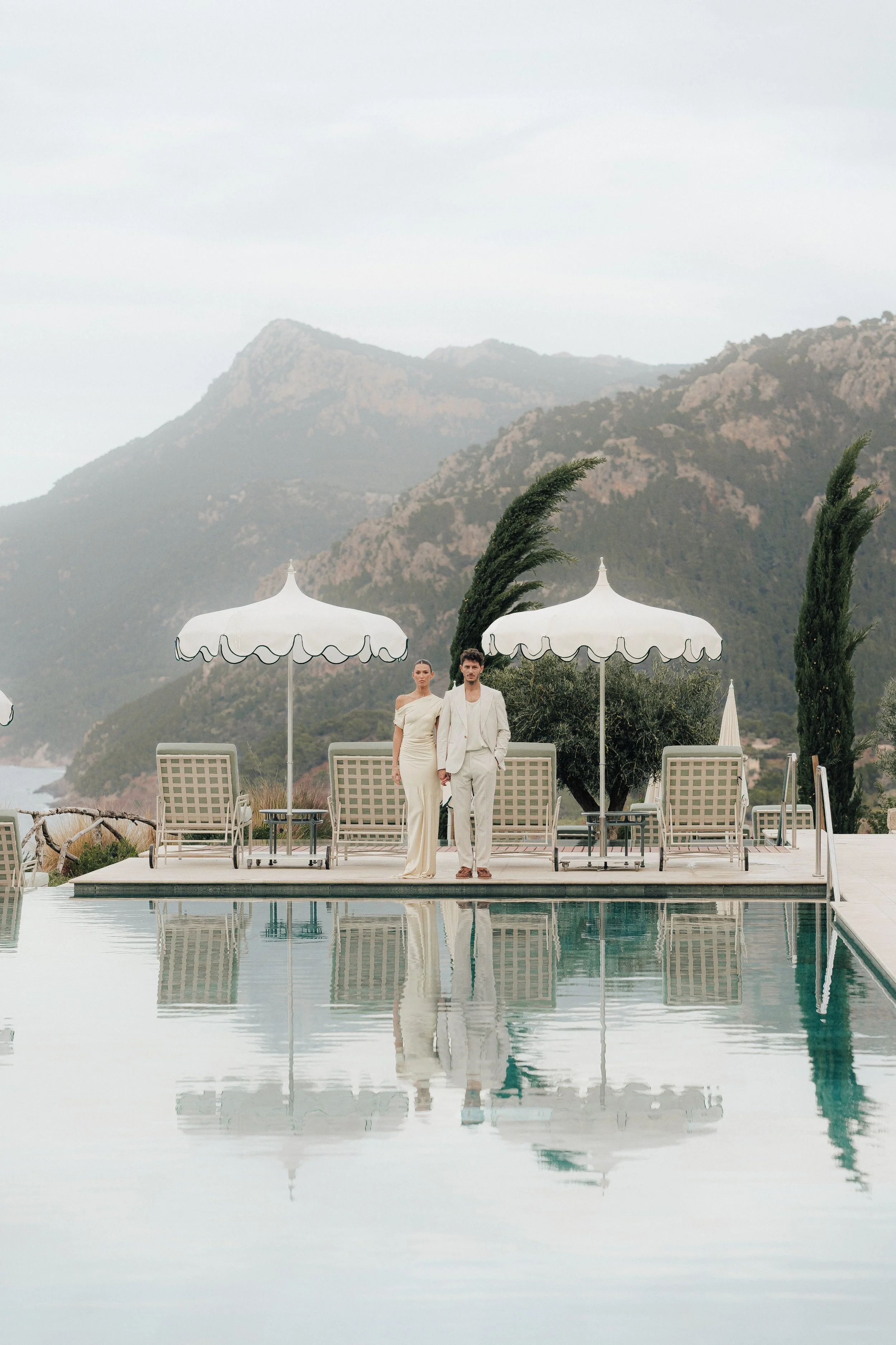 Ein Paar in weißen Kleidern steht an einem Pool vor einem Bergpanorama mit Windbäumen und Sonnenschirmen.