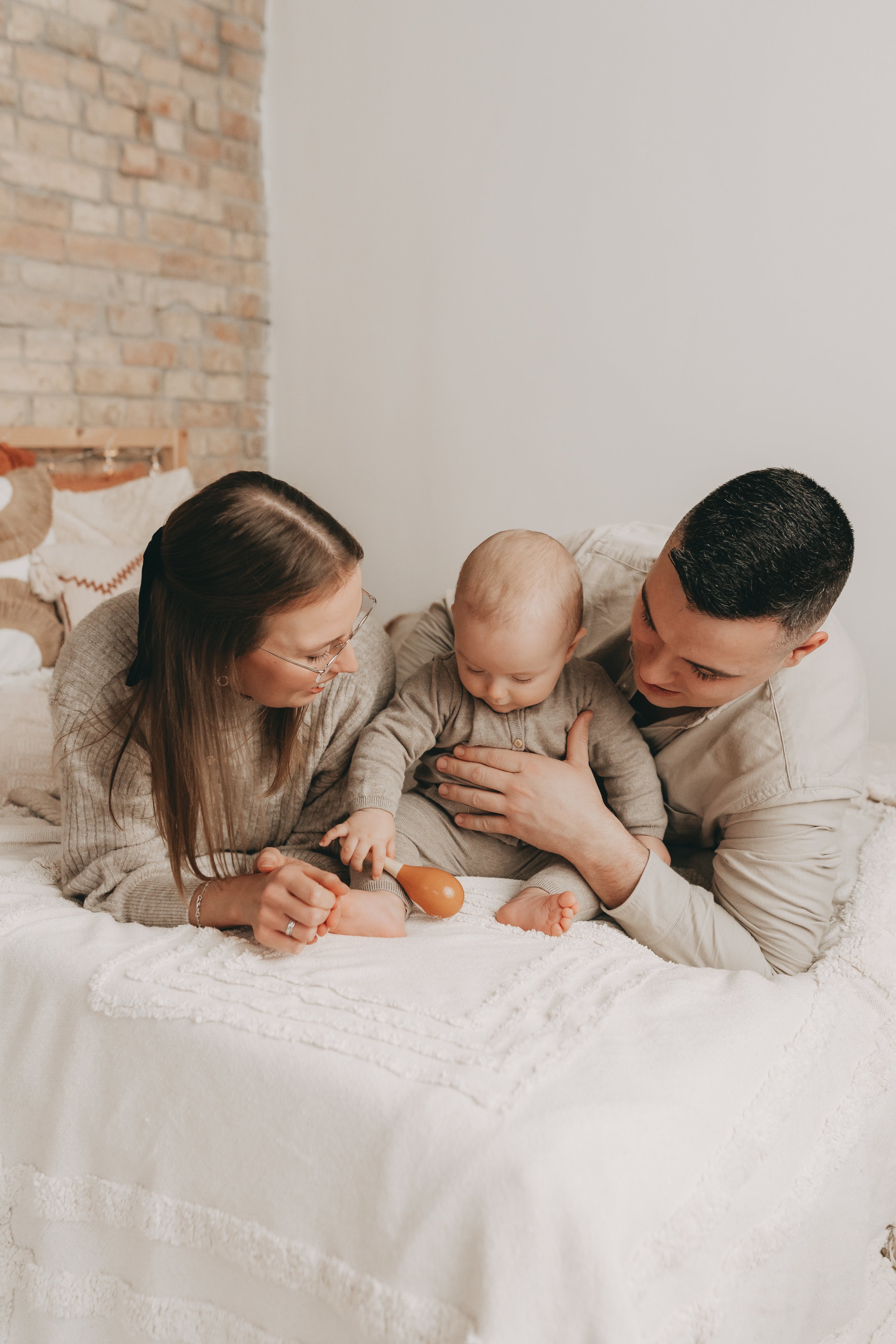 Eltern spielen mit ihrem Baby auf einem Bett. Das Baby hält ein Spielzeug in der Hand. Alle tragen bequeme Kleidung in einem gemütlichen, hellen Raum mit Backsteinmauer im Hintergrund.