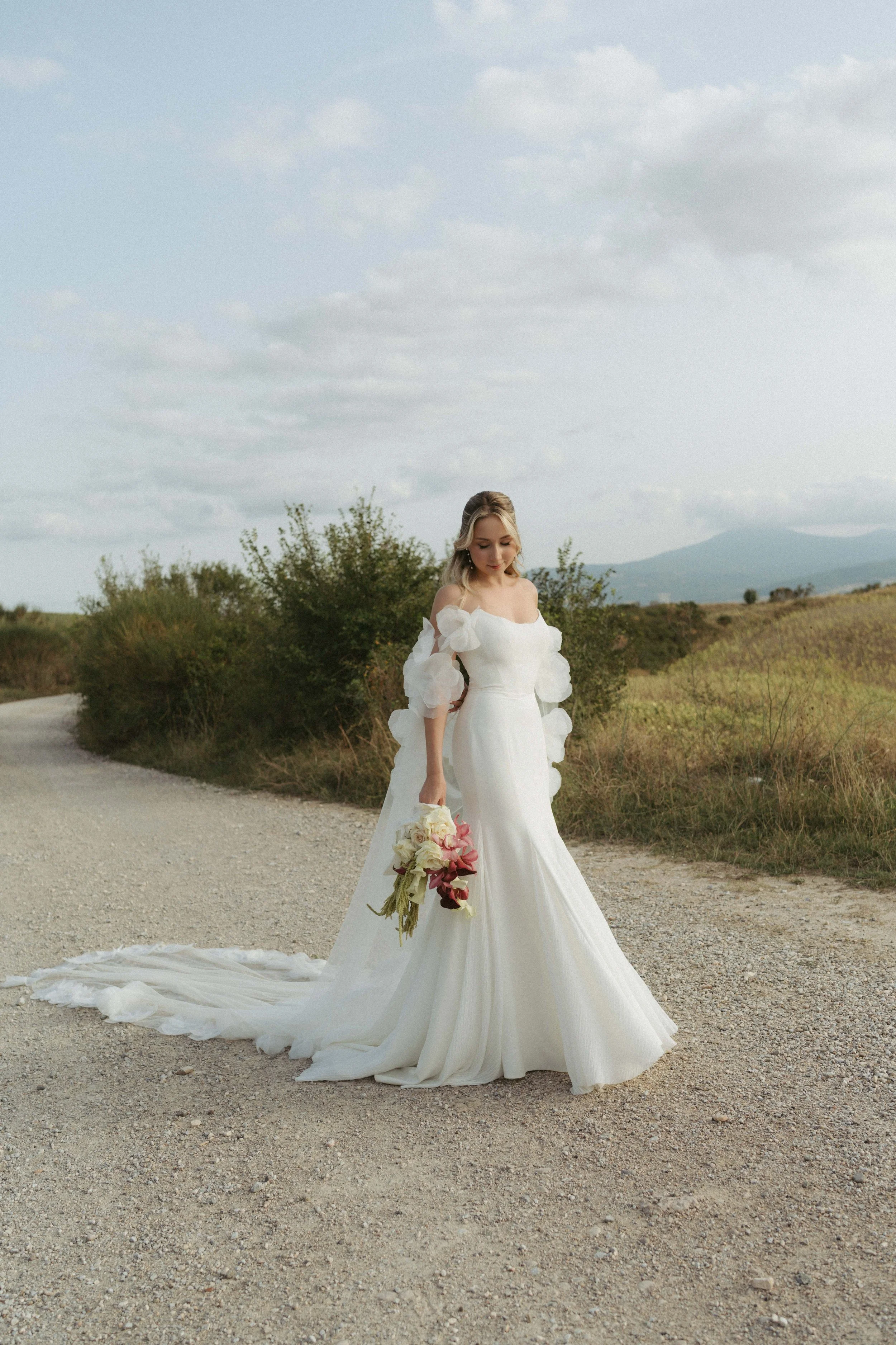 Frau in einem weißen Hochzeitskleid mit Blumenstrauß steht auf einem unbefestigten Weg in einer ländlichen Landschaft mit Hügeln im Hintergrund und einem bewölkten Himmel.