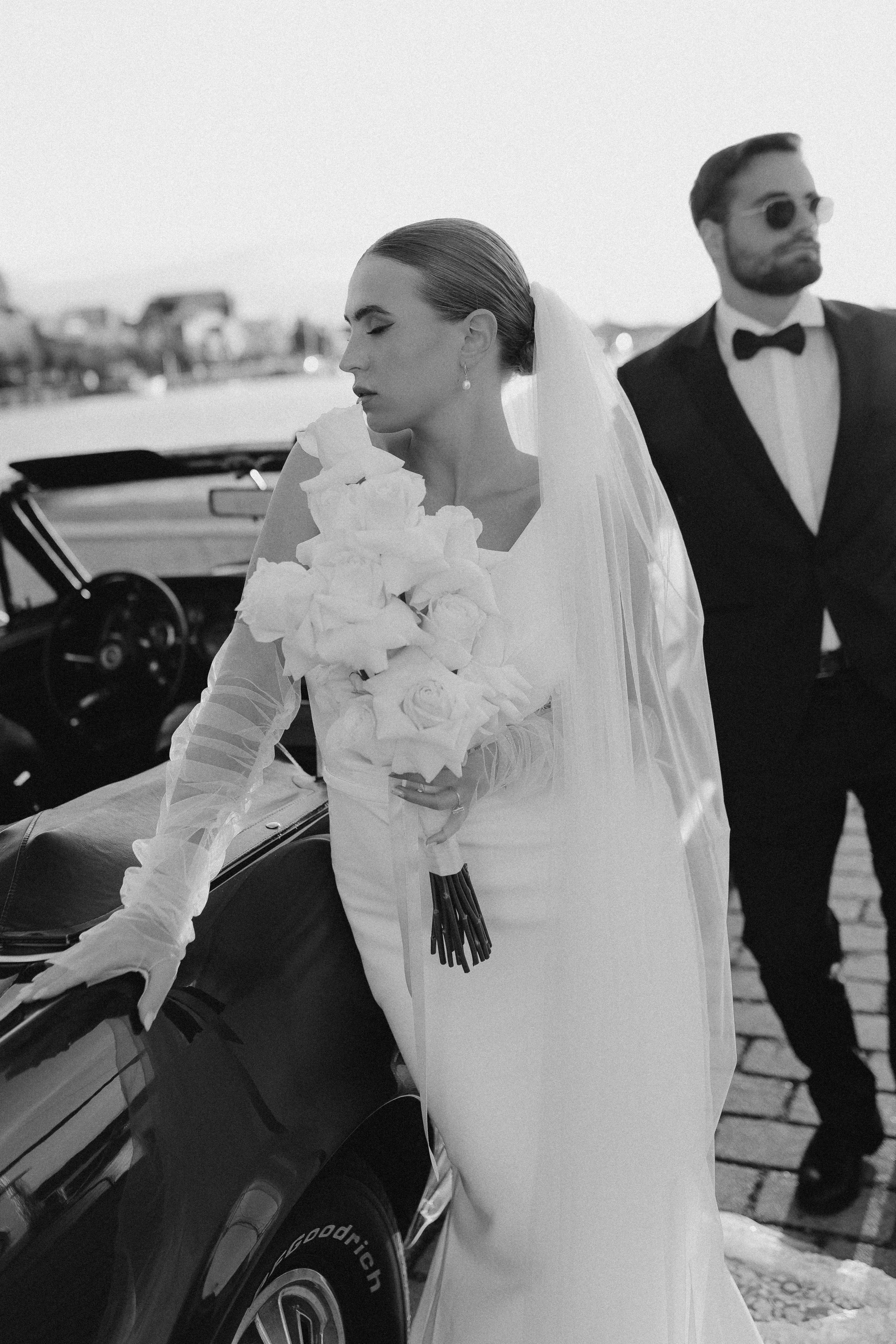 Braut in Hochzeitskleid mit Blumenstrauß neben einem schwarzen Oldtimer, im Hintergrund eine Stadt am Wasser, Mann in Smoking mit Sonnenbrille